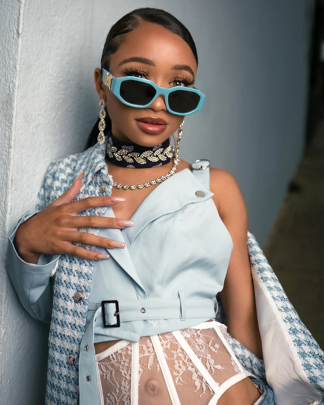 A woman wearing large, light blue sunglasses, pearl jewelry, a patterned neck scarf, and a stylish vest over a sheer lace top, leaning against a wall.