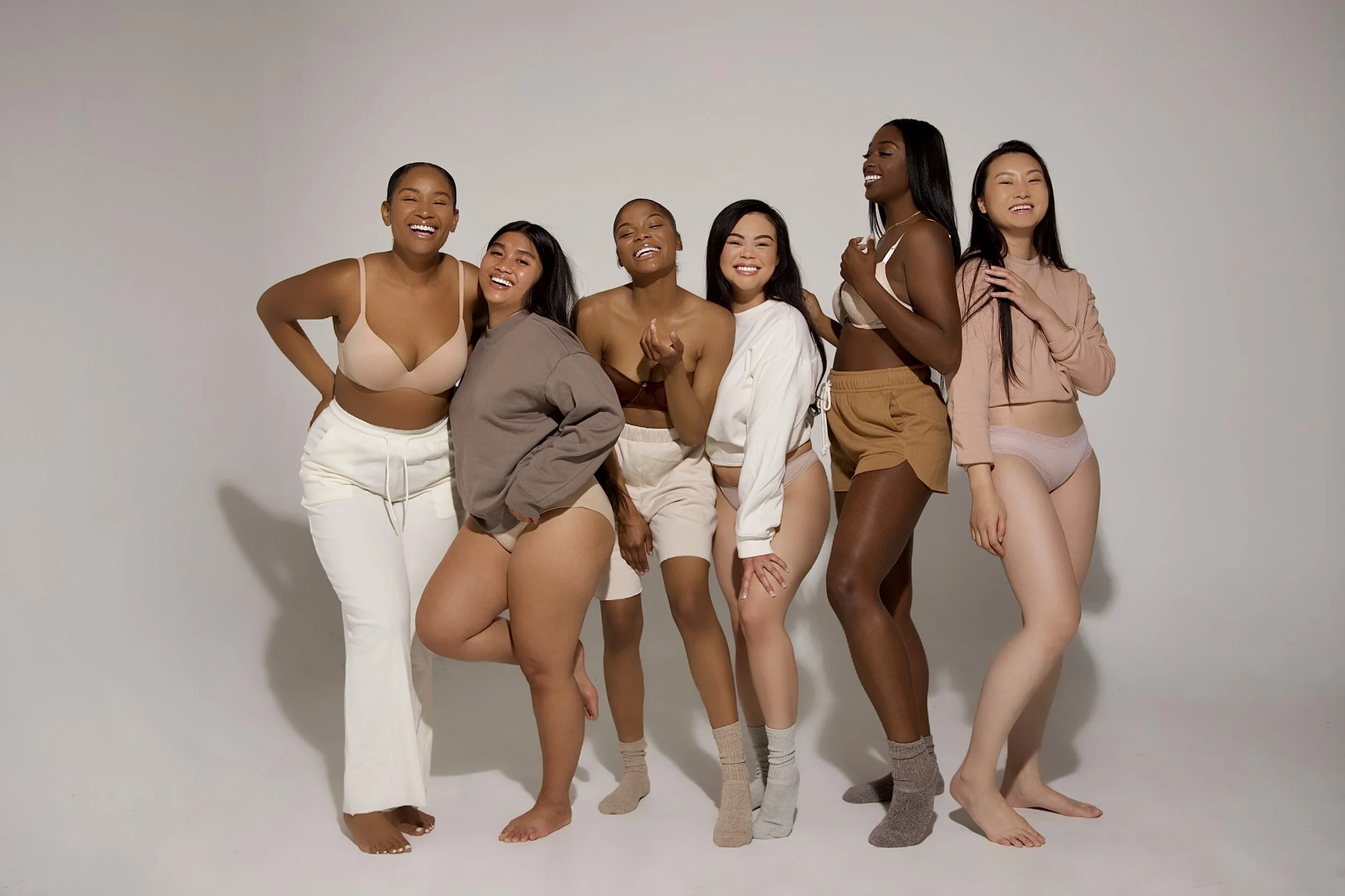 Group of seven women with diverse skin tones, smiling and standing together against a plain white background, dressed in casual and loungewear clothing.