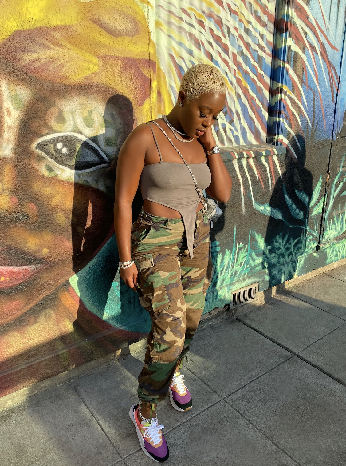 A woman with short blonde hair, wearing a beige crop top with cutouts, camouflage cargo pants, and colorful sneakers, standing against a vibrant mural wall.