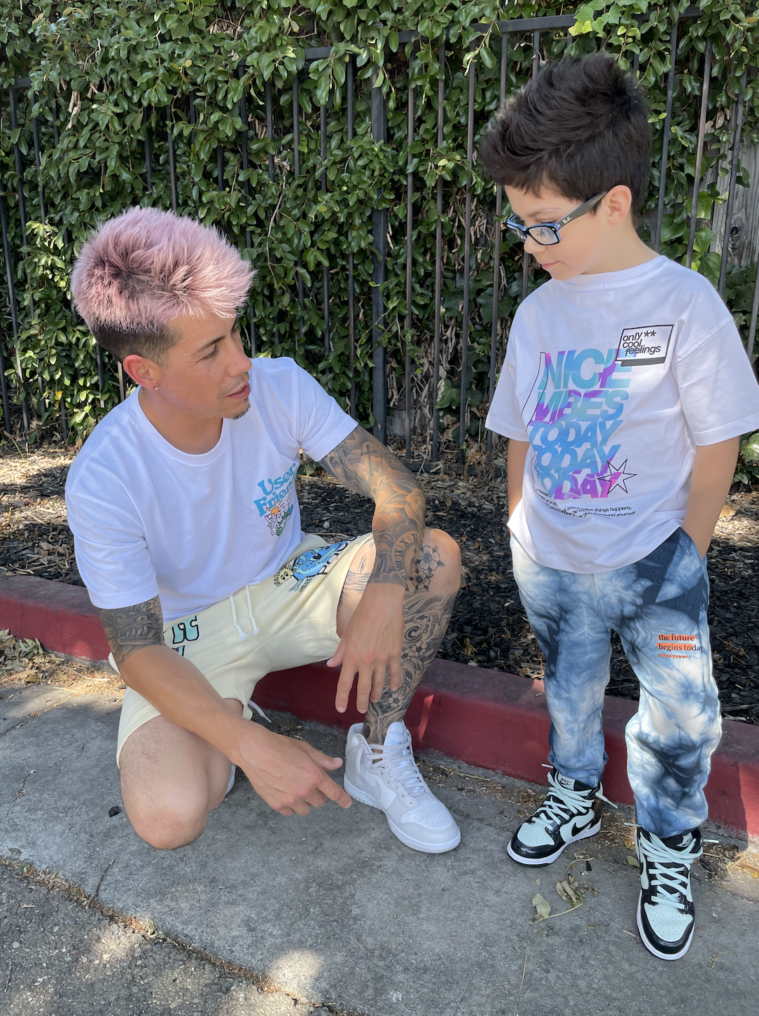 A man with pink hair and tattoos is squatting on the sidewalk, tying the shoelaces of a young boy with glasses. The man wears a white t-shirt and light-colored shorts, while the boy wears a white t-shirt with colorful text and tie-dye pants. They are