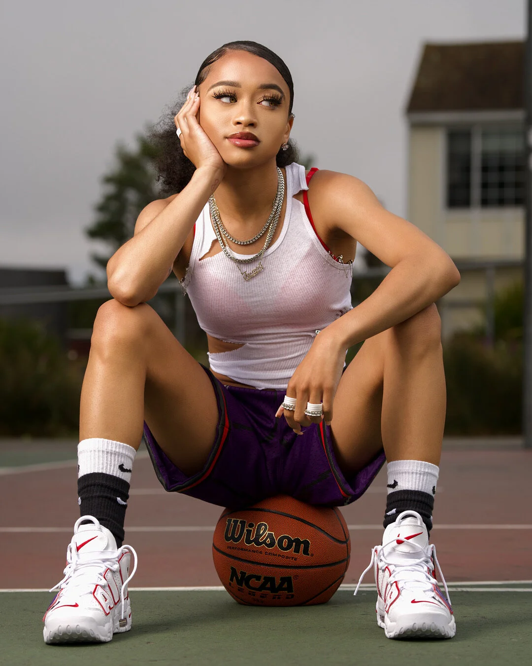 Young woman sitting on a basketball court with her hand on her face, wearing white sneakers, athletic shorts, a ripped white tank top, and jewelry, with a basketball between her legs.