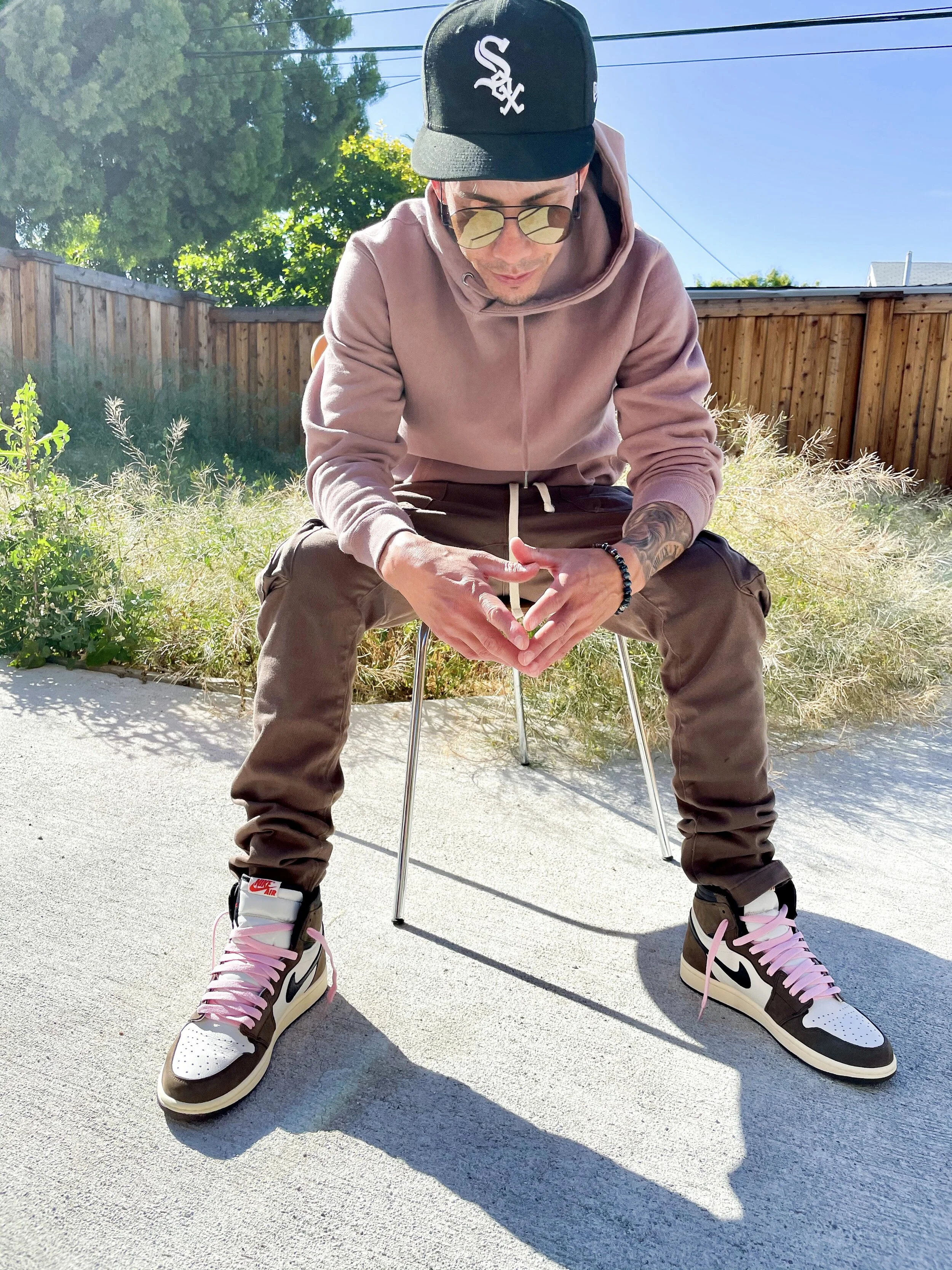 A young man wearing a black cap with a white logo, sunglasses, a pink hoodie, brown cargo pants, and Nike sneakers with pink laces, sitting on a metal chair outdoors on a sunny day with a wooden fence and greenery in the background.