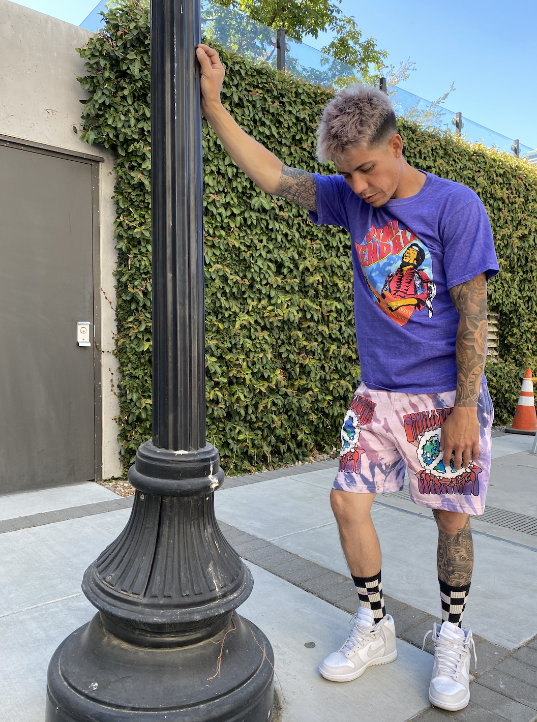 A young man with tattoos and gray hair wearing a colorful graphic t-shirt, pink shorts with bold prints, black and white checkered socks, and white sneakers, standing outdoors next to a black street lamp post, looking down.