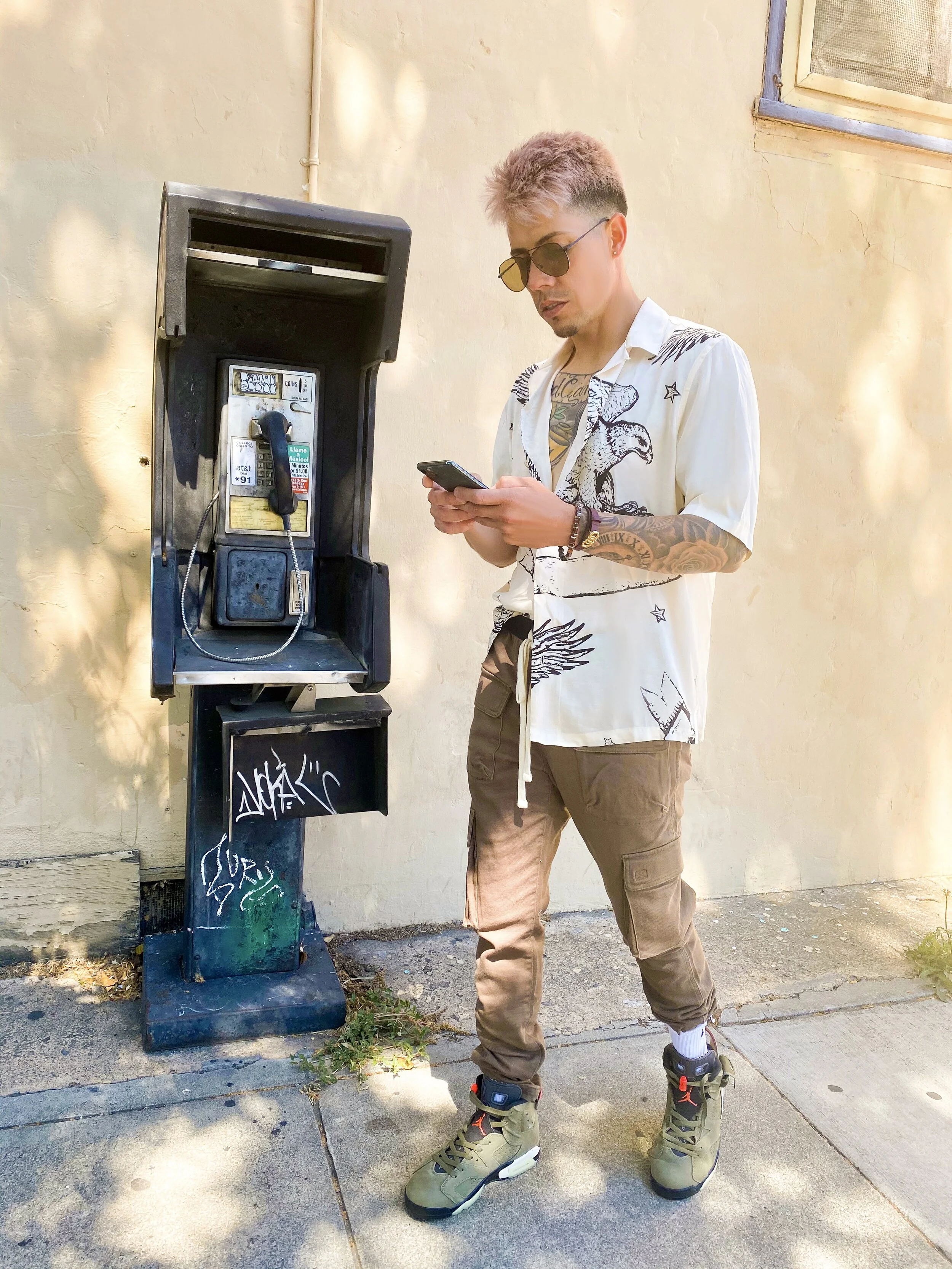 A young man with short, styled hair and sunglasses standing next to a payphone on a sidewalk, looking at his phone.