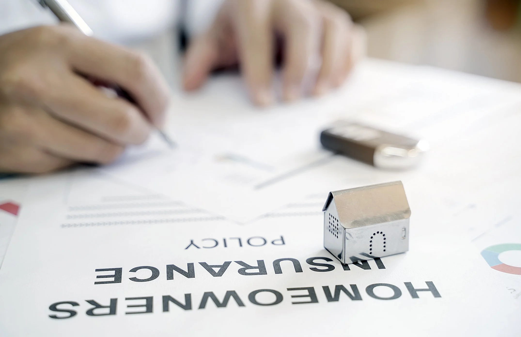 Person filling out homeowners insurance policy form with miniature house on desk.