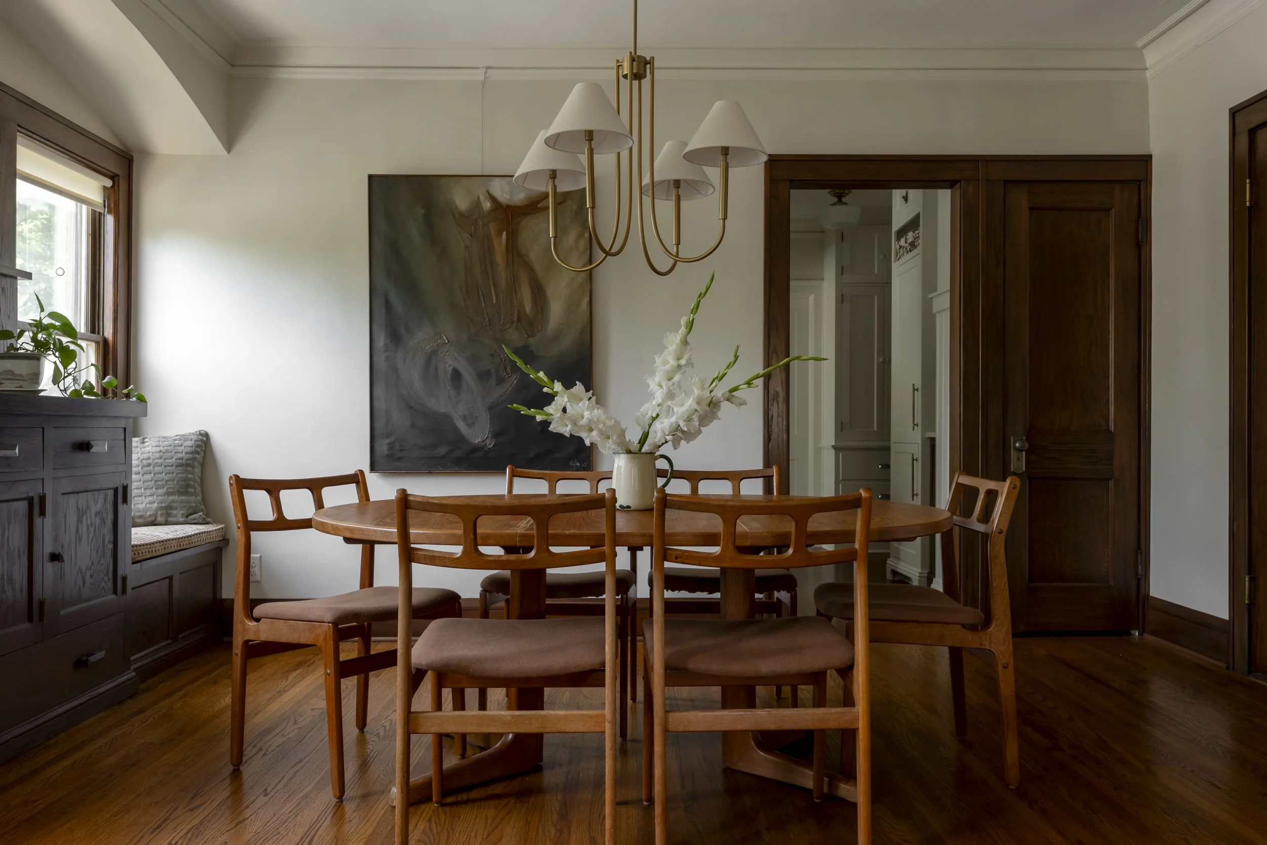 A wooden dining table with six matching chairs, a vase of white flowers, a chandelier overhead, and a dark abstract painting on the wall, in a well-lit dining room with hardwood floors.