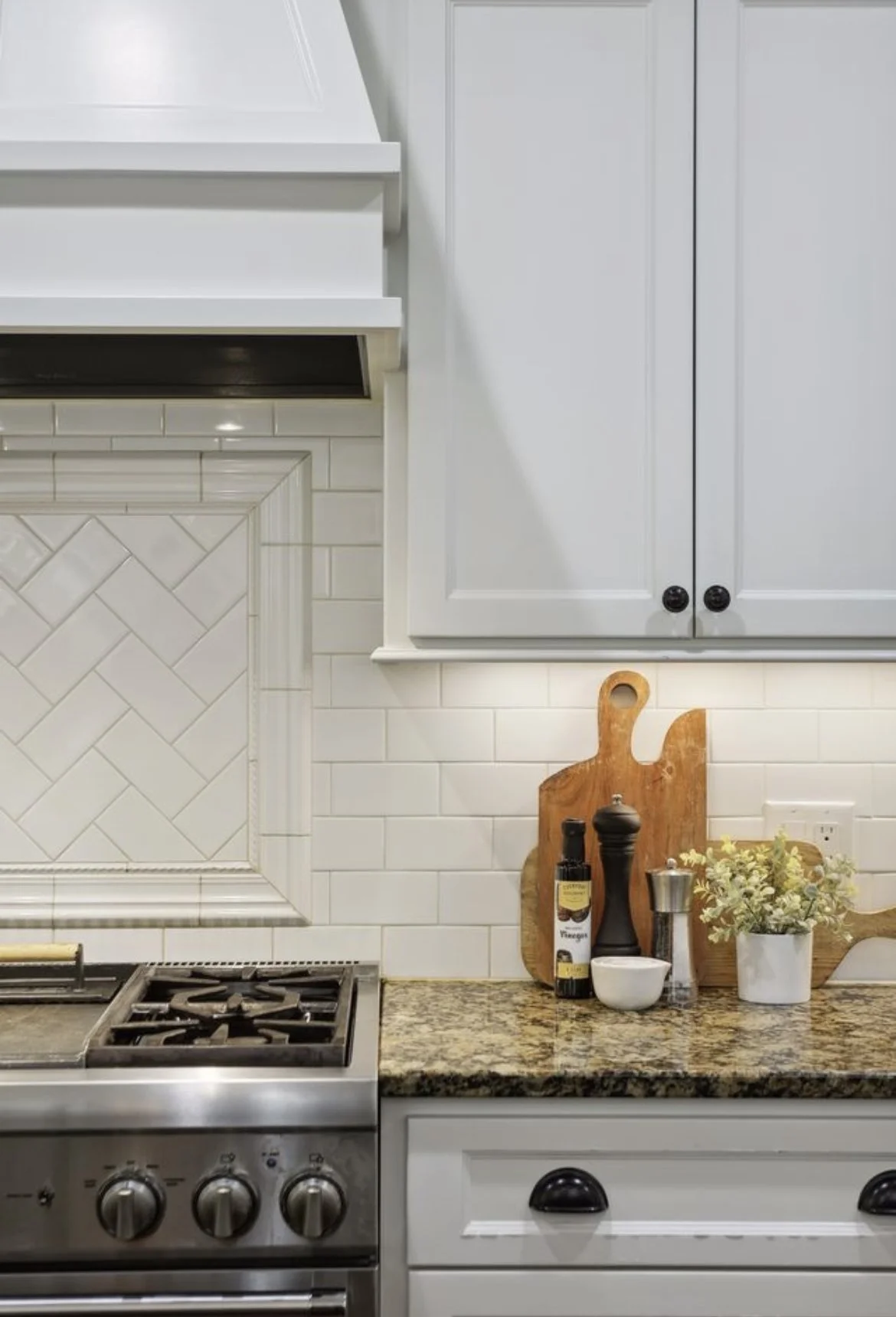A close-up view of a kitchen countertop with a stove, cutting boards, a small white bowl, a plant, and spice bottles, against a background of white tiled backsplash and light gray cabinets.