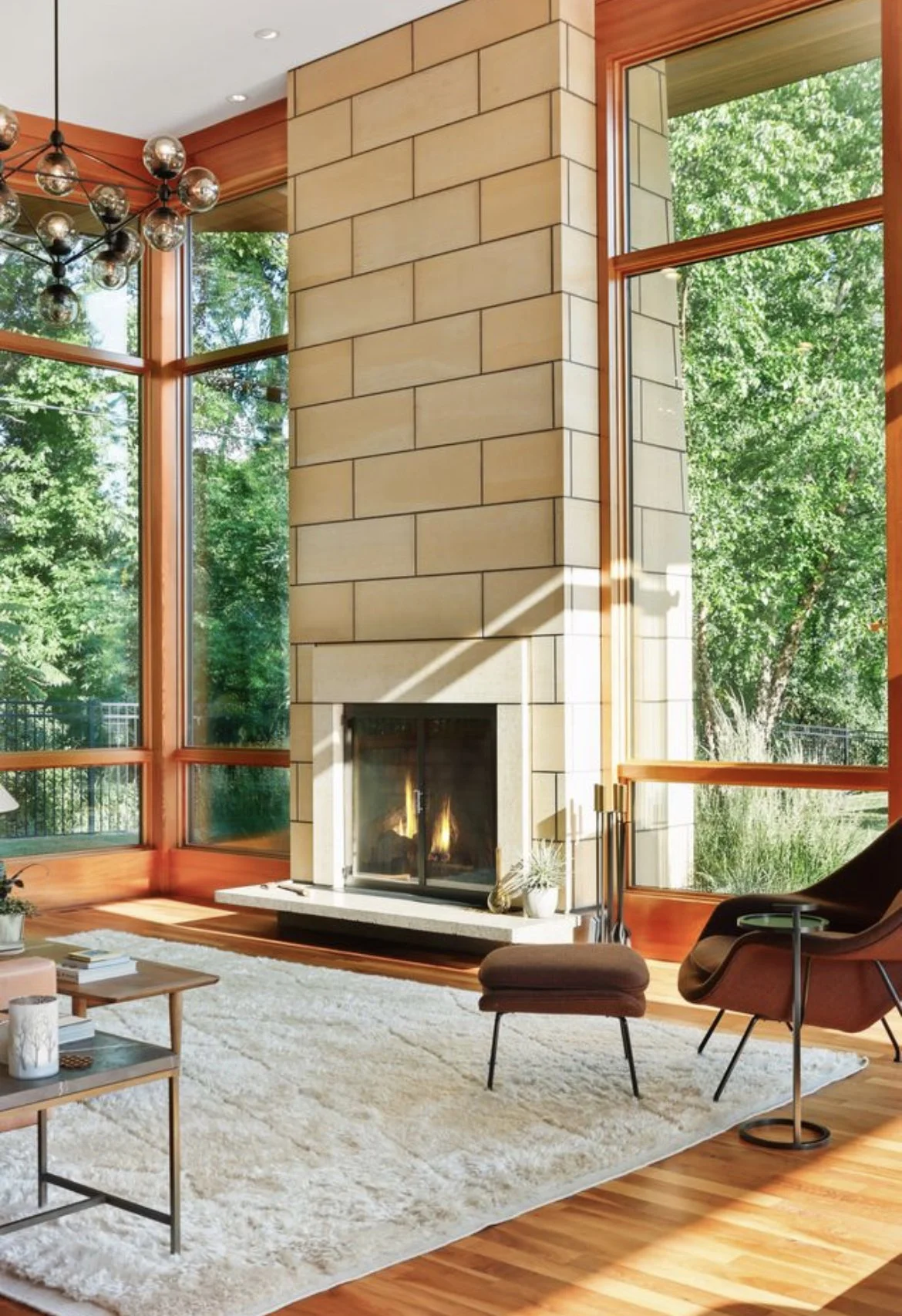 Living room with large windows, a stone fireplace with a fire burning, wooden floors, a modern chandelier, a black armchair with a small table, a beige rug, and decorative items near the fireplace.