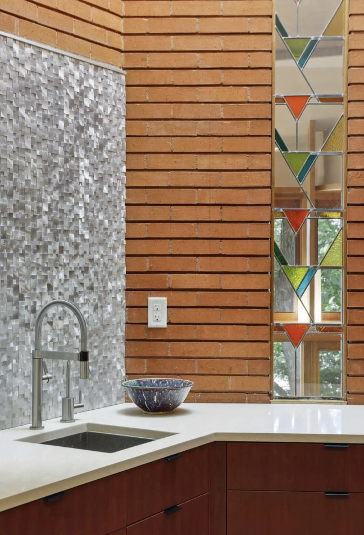 A kitchen corner with a white countertop, a black sink with a silver faucet, a decorative bowl, a textured gray wall, a brick wall, and a stained glass window with geometric shapes in orange, green, and yellow.