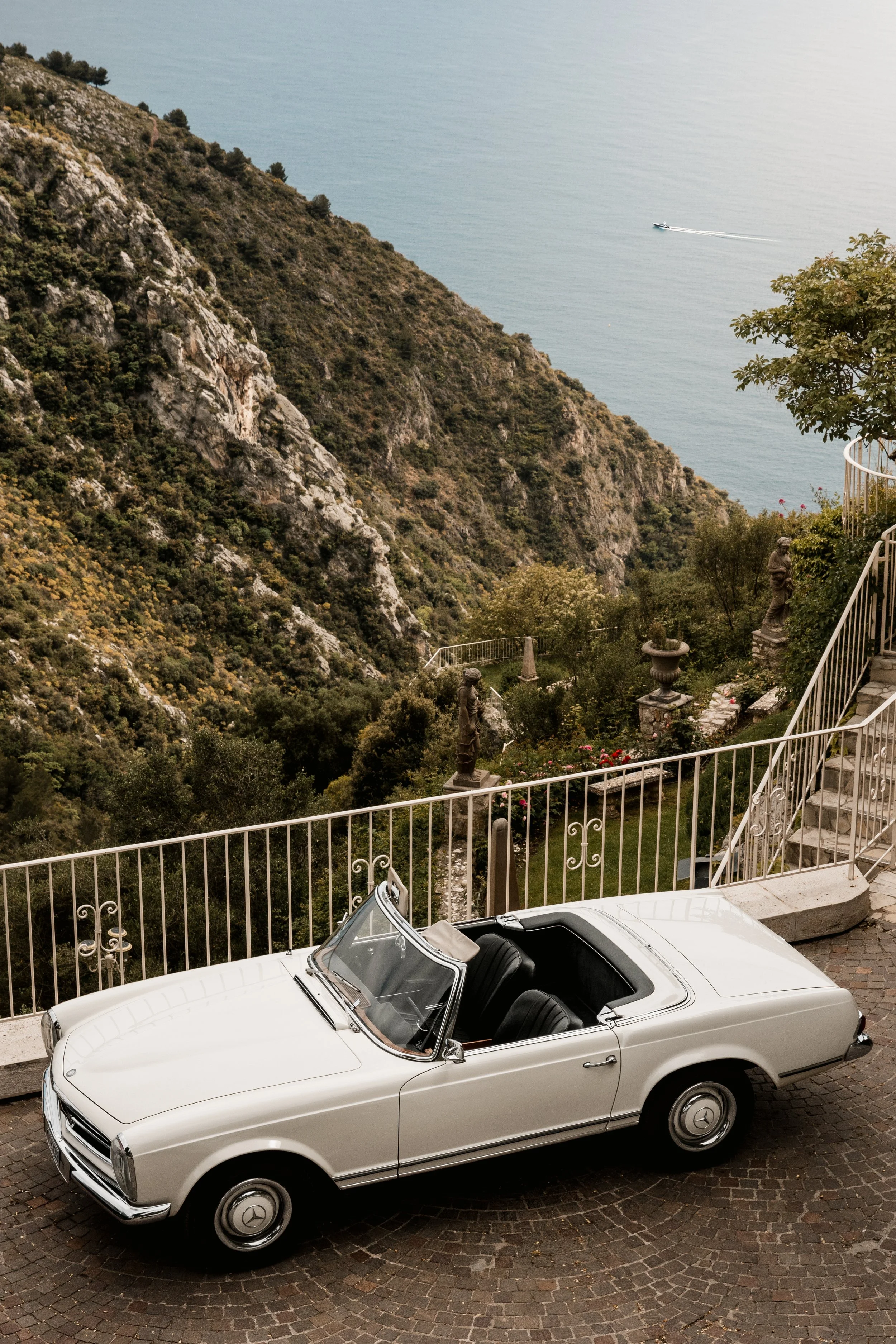 A white vintage convertible car parked on a cobblestone driveway overlooking a hillside with scenic garden decorations, a staircase, and an ocean in the background with a boat sailing.