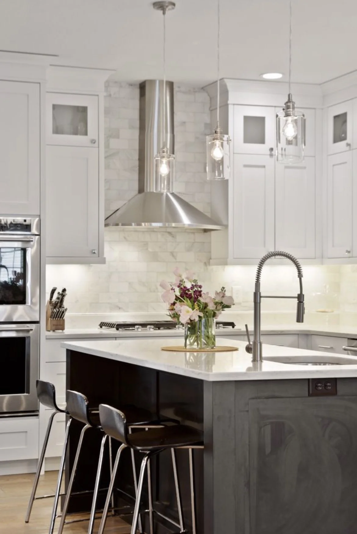 Modern kitchen with white cabinets, stainless steel oven, vent hood, and island with black base and three black chairs. There is a vase of pink and purple flowers on the island and pendant lights hanging from the ceiling.