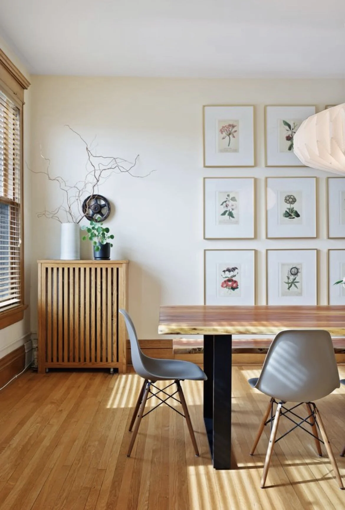 A dining room with wooden flooring, a wooden table, two modern chairs, wall art with framed botanical prints, a wooden cabinet with plants and a decorative object, and a window with blinds.