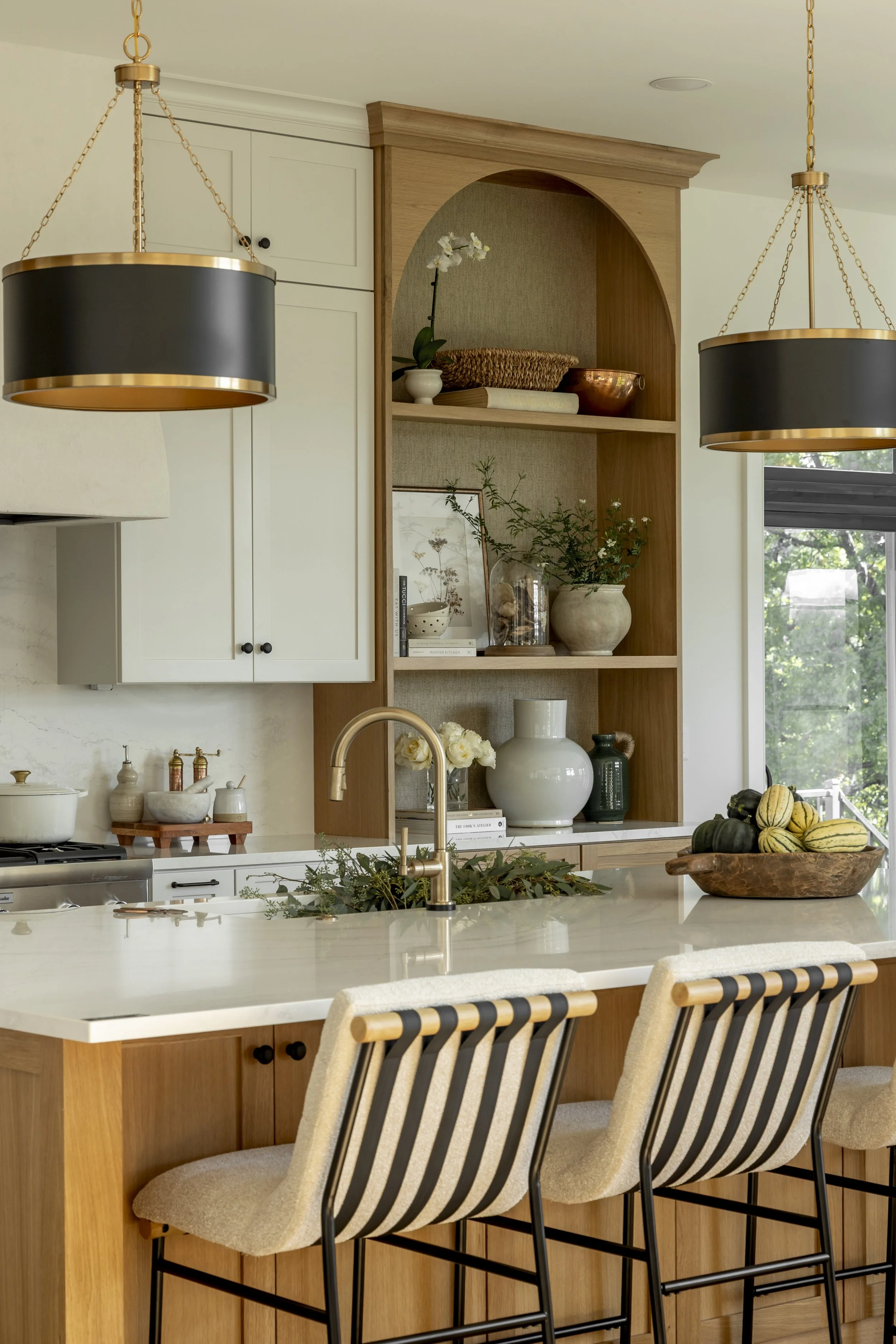 A modern kitchen with wooden and white cabinetry, black and gold pendant lights, an island with a marble countertop, barstools with beige cushions and black striped backs, open shelves with vases, plants, and decorative objects, a bowl of gourds, and