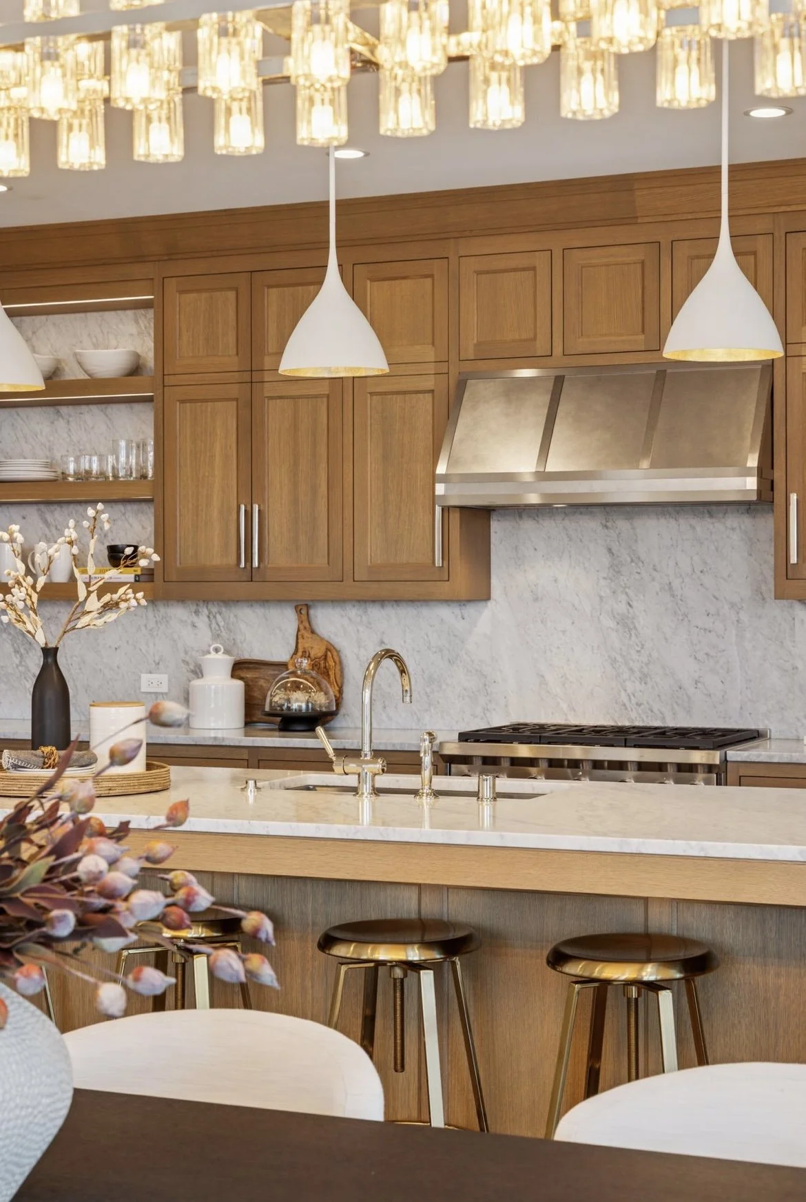 Modern kitchen with wooden cabinets, marble backsplash, and a white marble countertop island with two black and gold bar stools. White pendant lights hang from the ceiling, and open shelves display bowls, glasses, and decorative items. A vase with co