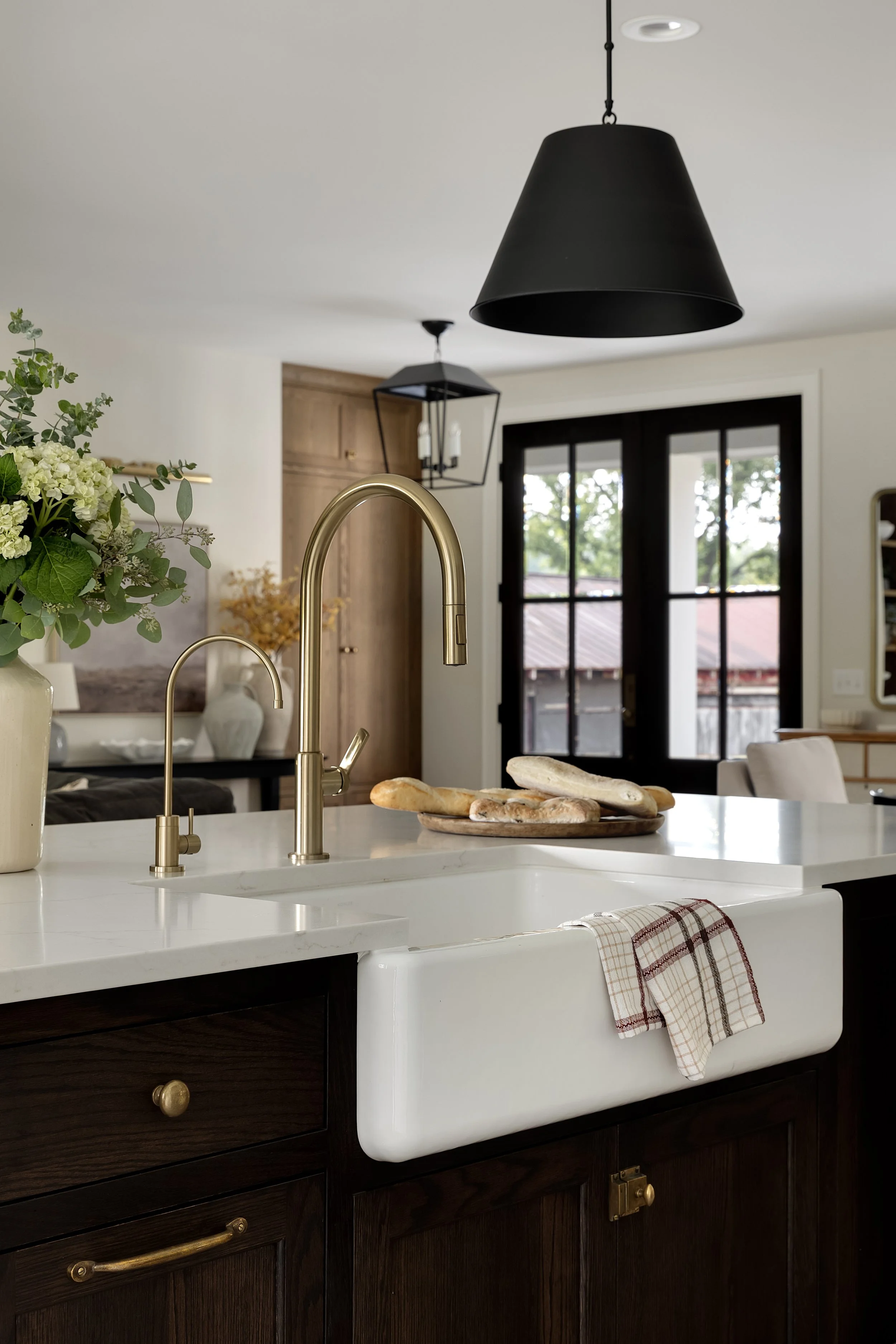 A modern kitchen with brass faucet, white farmhouse sink, and dark wooden cabinets. There are bread rolls on a wooden tray on the white marble countertop, a dish towel hanging from the sink, and a plant with white flowers in a beige vase. In the back