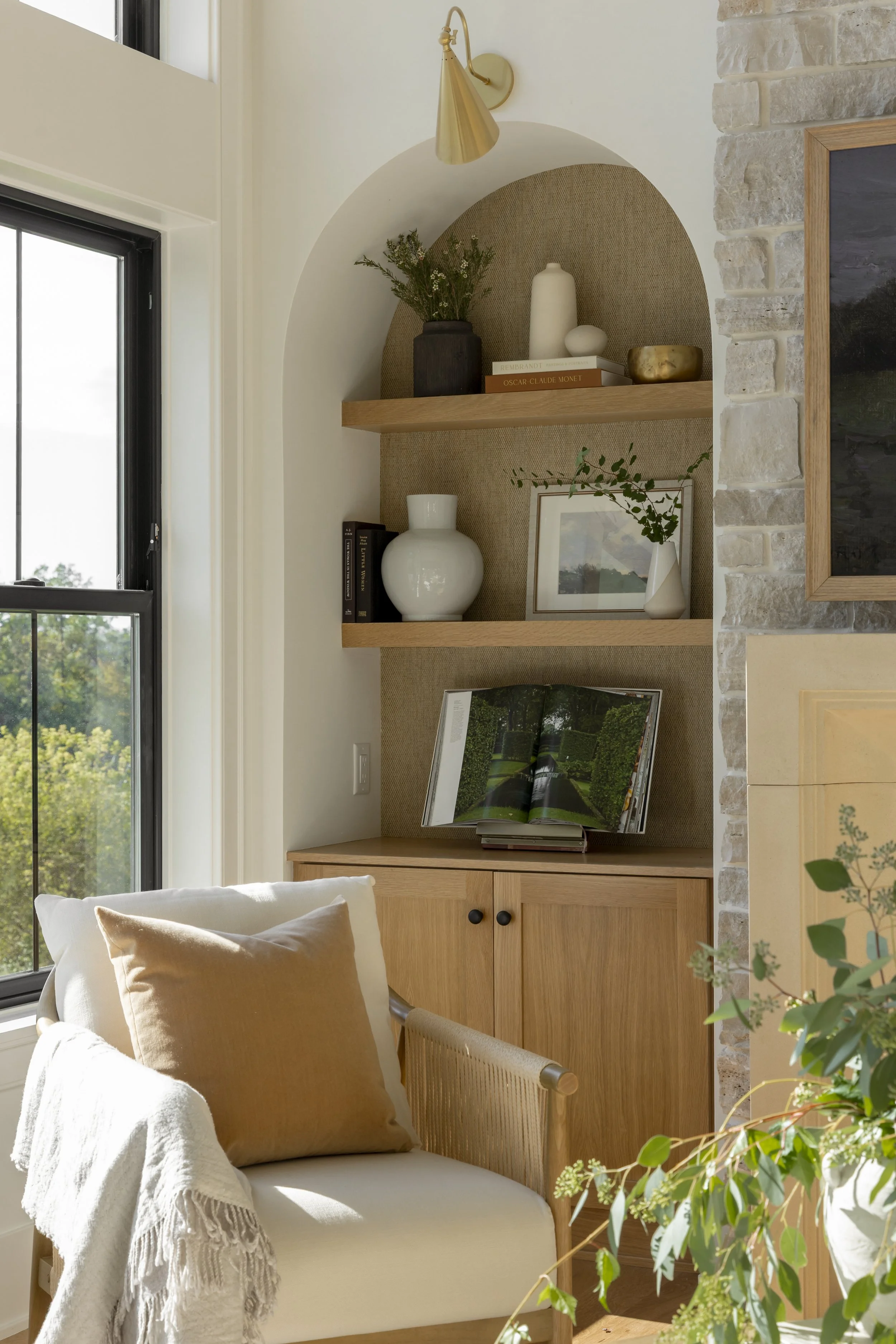 A cozy corner of a living room with a white armchair, beige pillow, and a white throw blanket. Behind the armchair is a built-in wooden cabinet with open shelves containing decorative vases, books, and framed artwork. To the left, there's a window wi