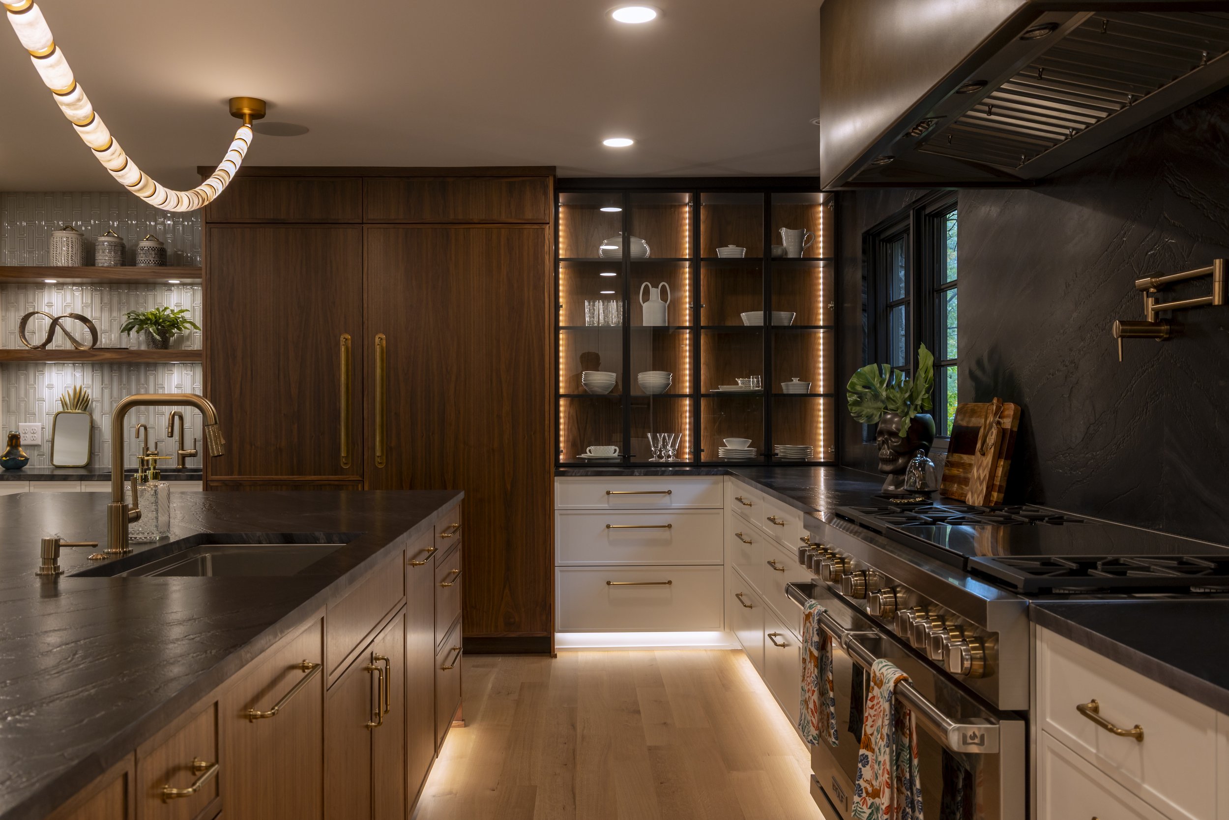 Modern kitchen with dark countertops, white and wood cabinets, gold hardware, open shelving, and decorative lighting.