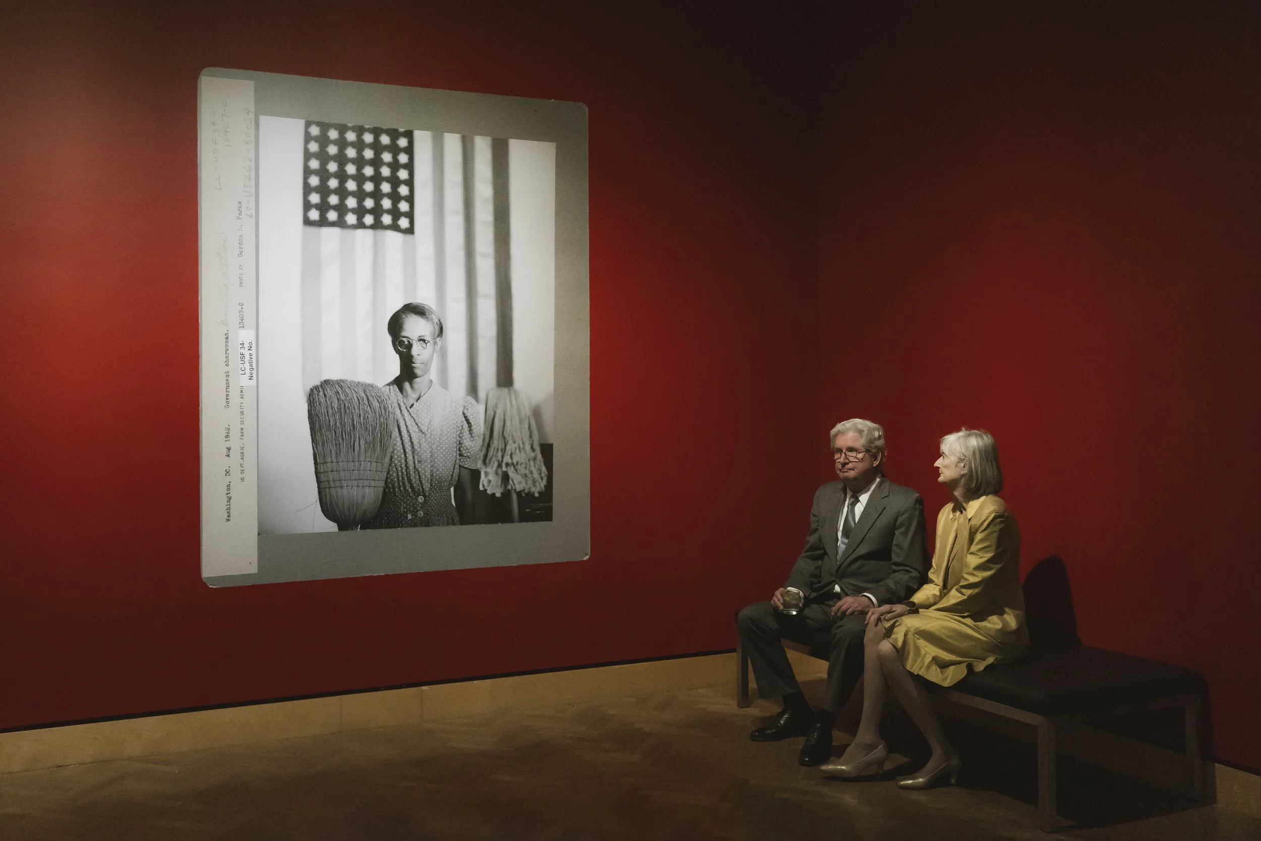 An art exhibit with a large black and white photo of a woman with short hair and glasses, standing in front of an American flag, displayed on a red wall. Two elderly people, a man in a suit and tie holding a glass and a woman in a yellow dress, sit o