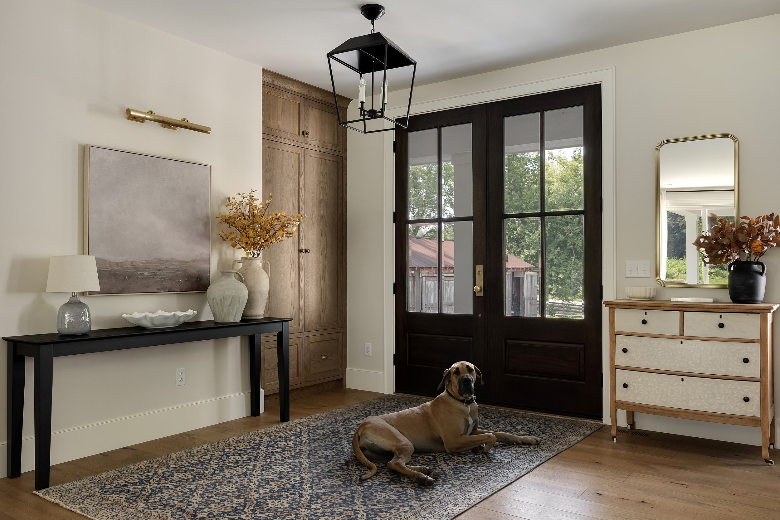 Living room with wooden flooring, a black front door with glass panels, and a large dog lying on a patterned rug. There is a black console table with vases and a lamp on it, a wooden cabinet with drawers, a mirror, and artwork on the walls.