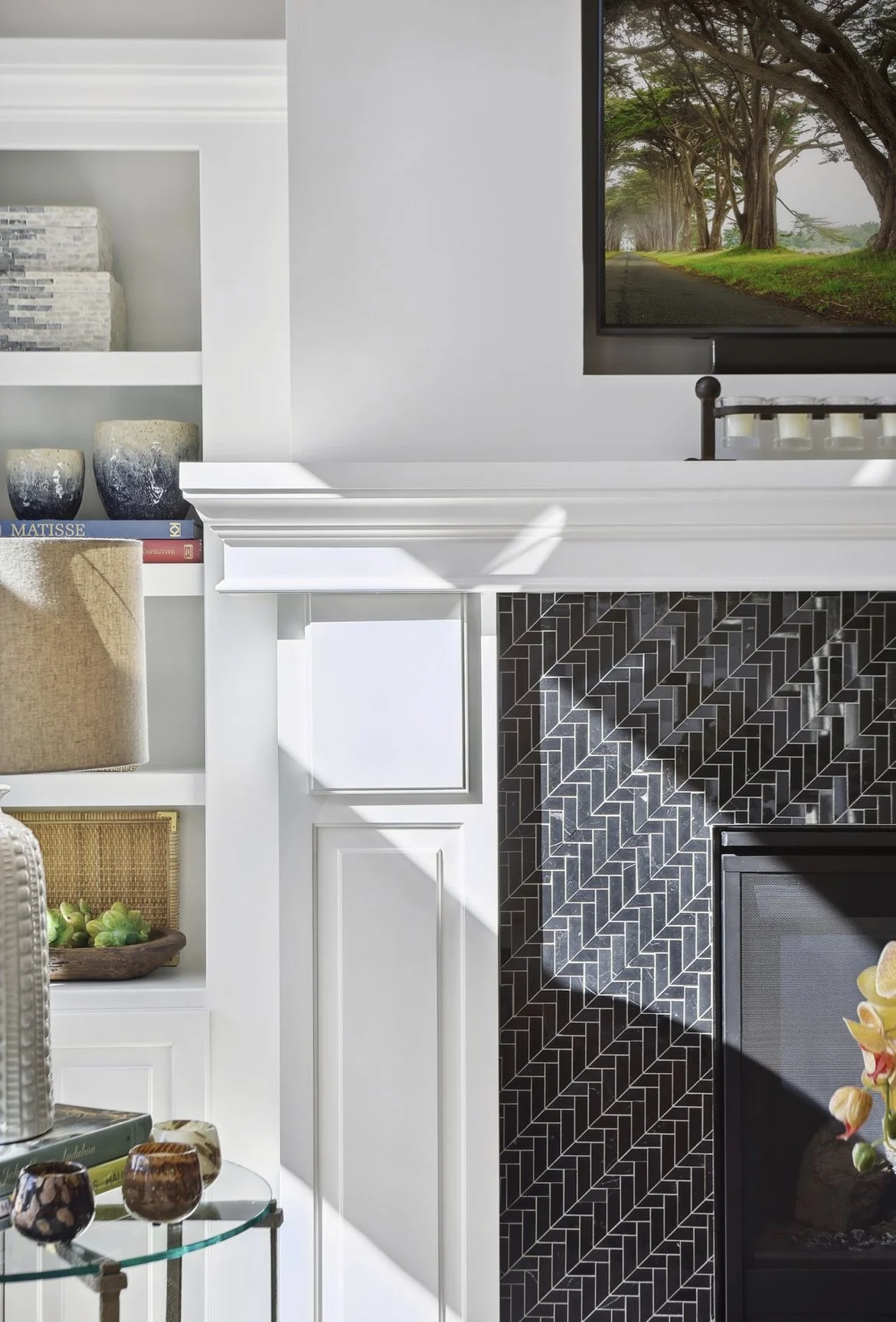 Close-up of a living room corner with a white fireplace mantle and black herringbone tile surround, a mounted flat-screen TV displaying a tree-lined road, and built-in shelves with decorative items and books.