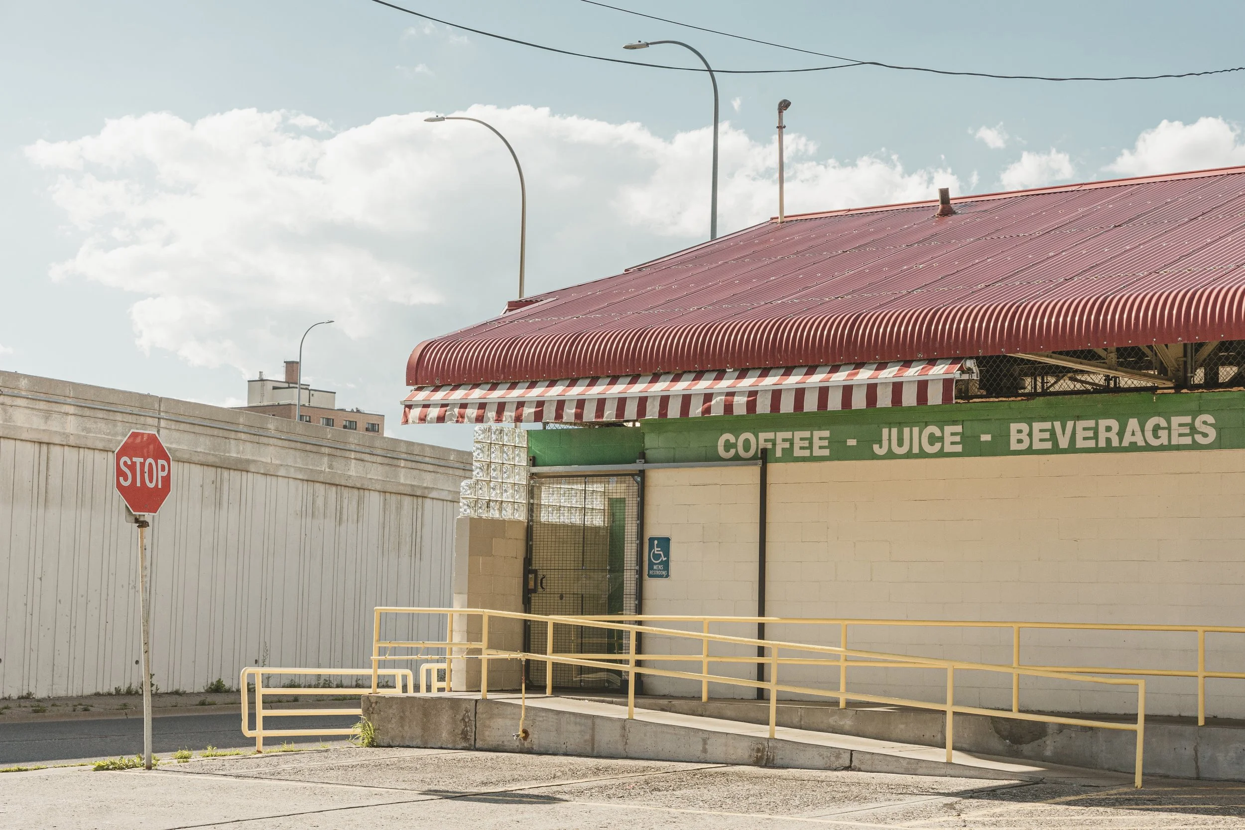 A small building with a red, corrugated metal roof and an awning, with a sign indicating it sells coffee, juice, and beverages. There is a handicap accessible ramp and a stop sign in the foreground, with a white fence, street, and cloudy sky in the b