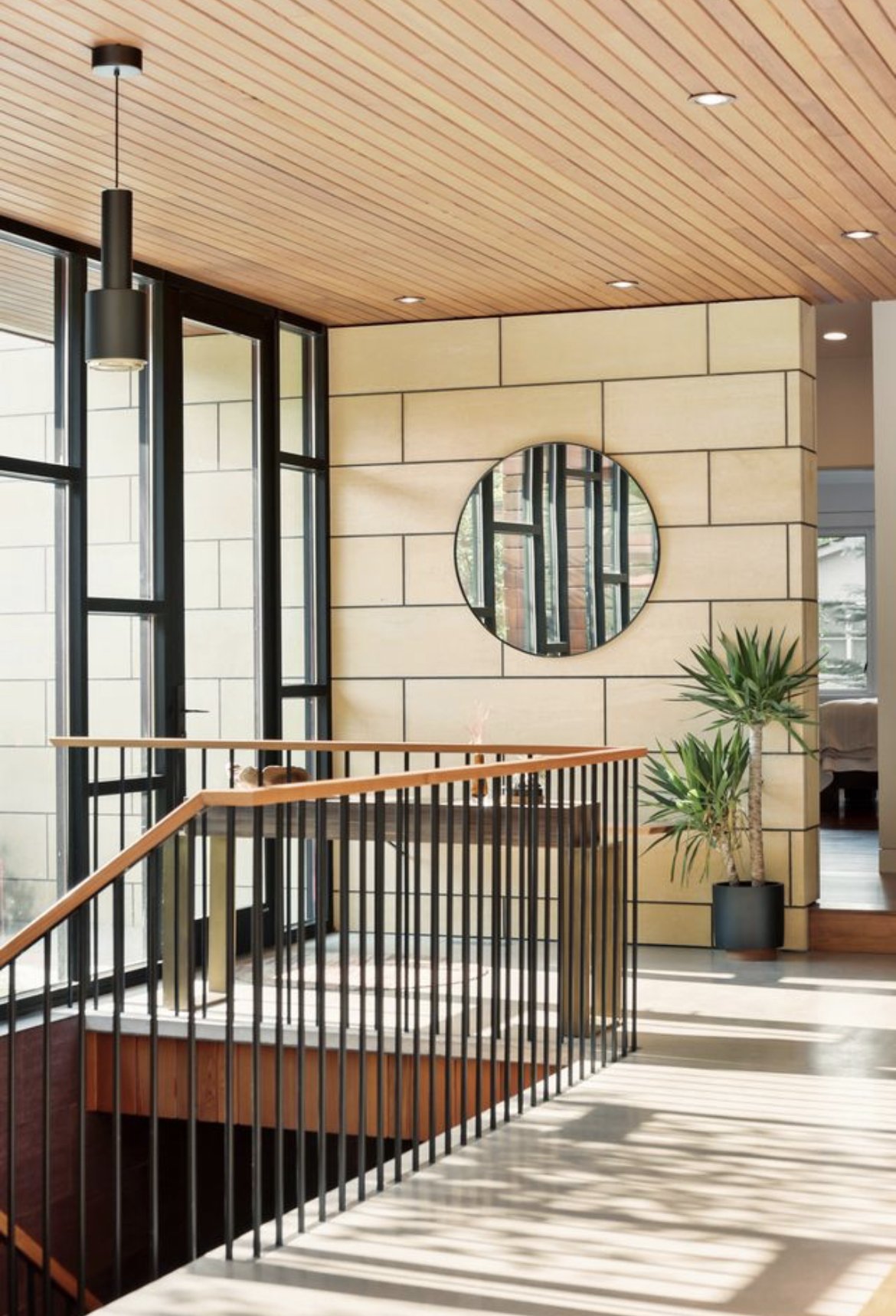 Modern interior space with large windows, wood ceiling, interior plants, and a round mirror on a beige tiled wall.