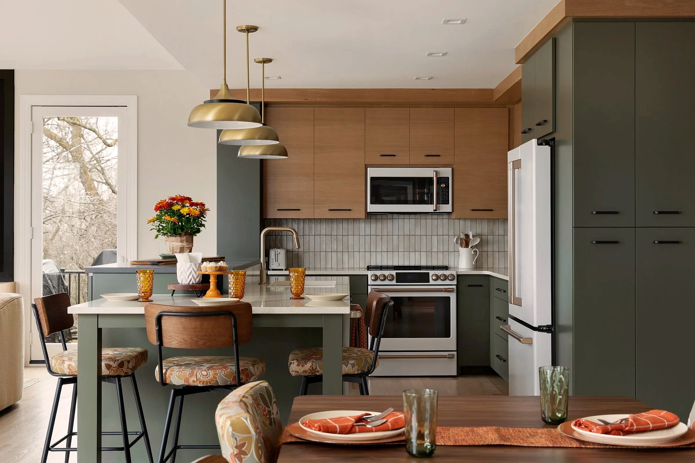 Modern kitchen with green and wood cabinets, white backsplash, stainless steel appliances, and a dining area with a wooden table set for two