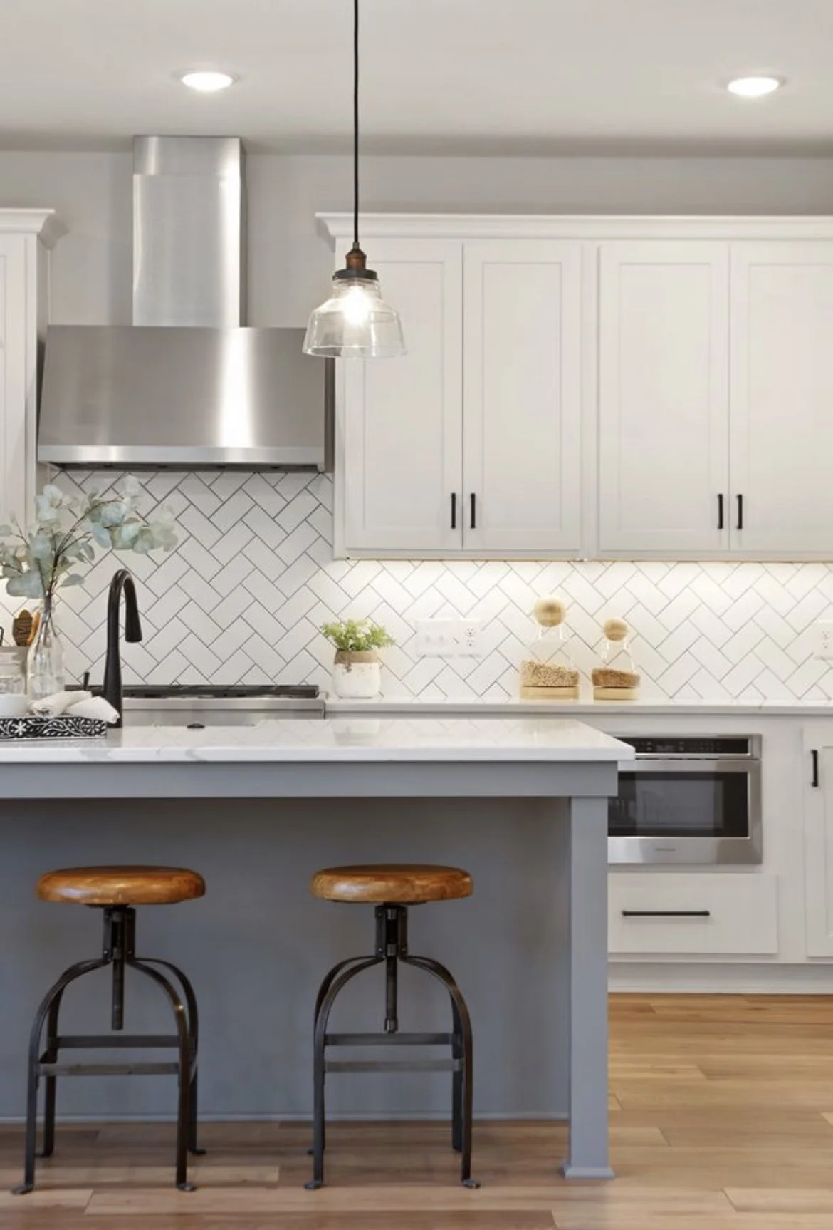 Modern kitchen with white cabinets, stainless steel appliances, a white subway tile backsplash, and a gray kitchen island with two rustic wooden stools.