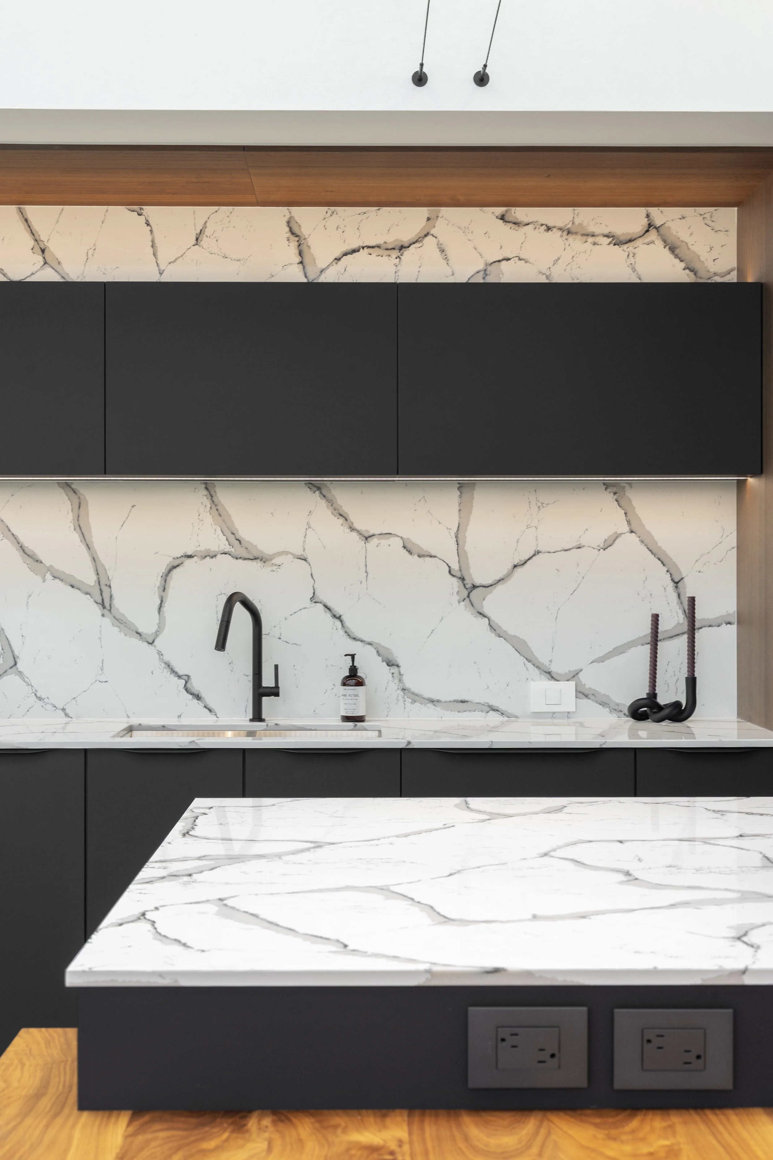 Modern kitchen with black cabinetry, white marble countertops and backsplash with dark gray veins, black faucet, a soap dispenser, and decorative black hooks.