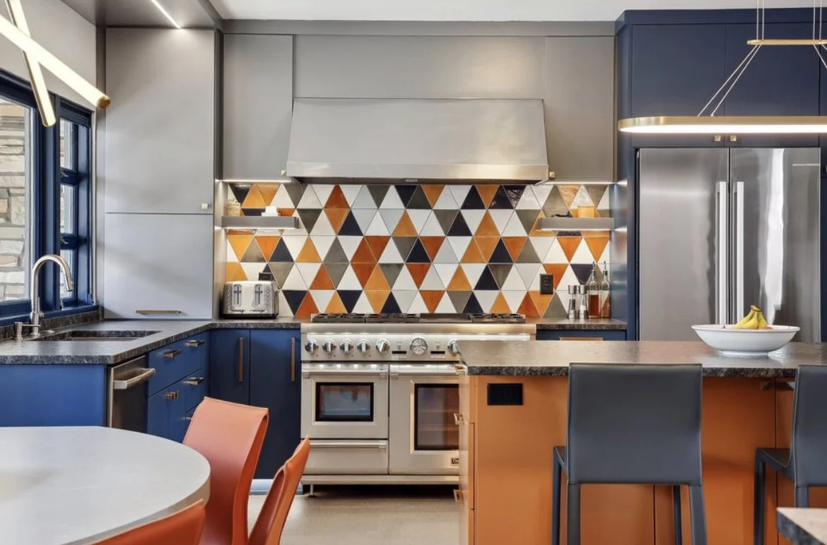 Modern kitchen with navy blue cabinets, geometric patterned backsplash in orange, black, and white, stainless steel appliances, and a white bowl of bananas on an island counter.
