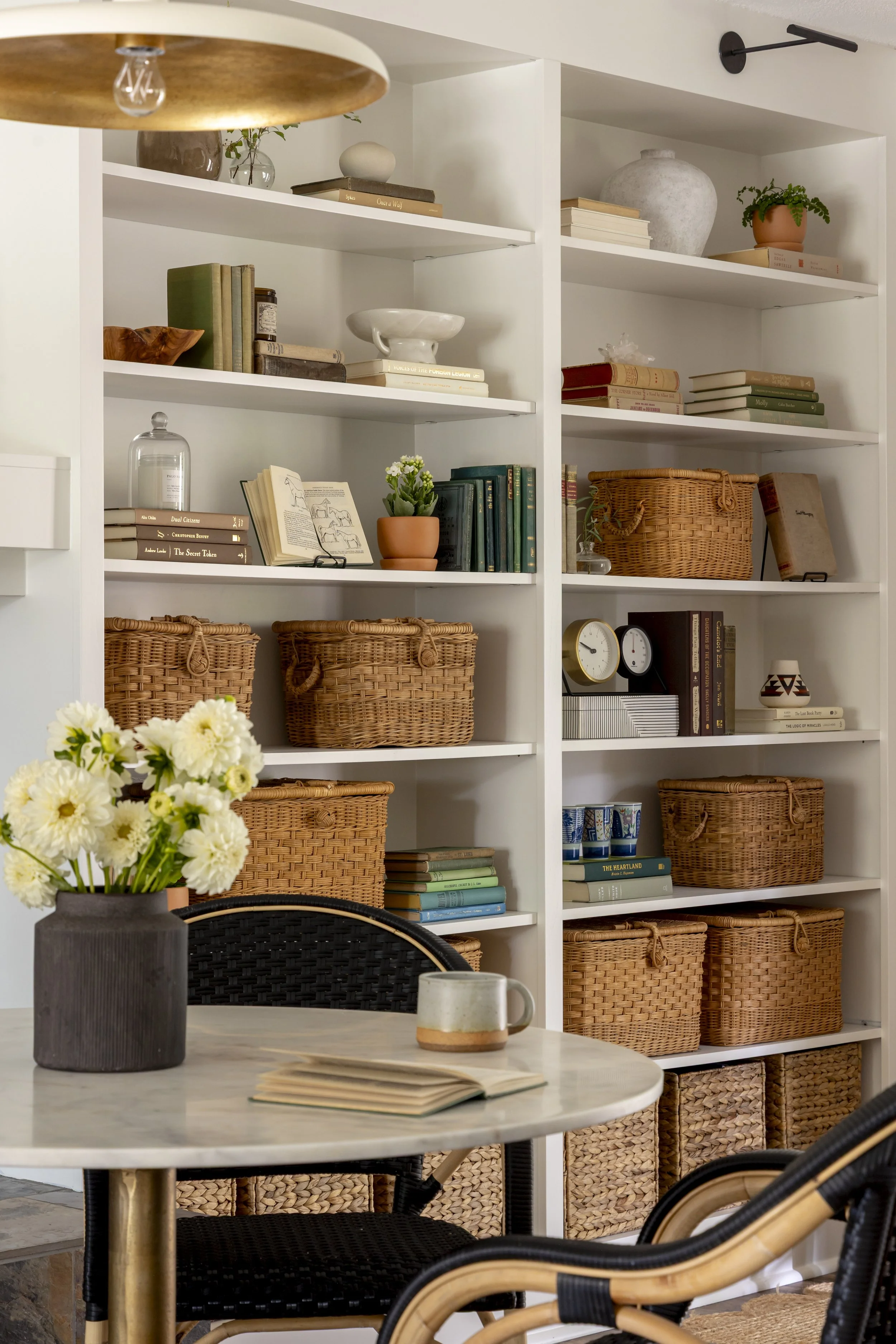A white bookshelf filled with books, wicker baskets, decorative items, and potted plants. In front, a round marble table with an open book, a mug, and a flower vase, surrounded by black woven chairs with rattan accents.