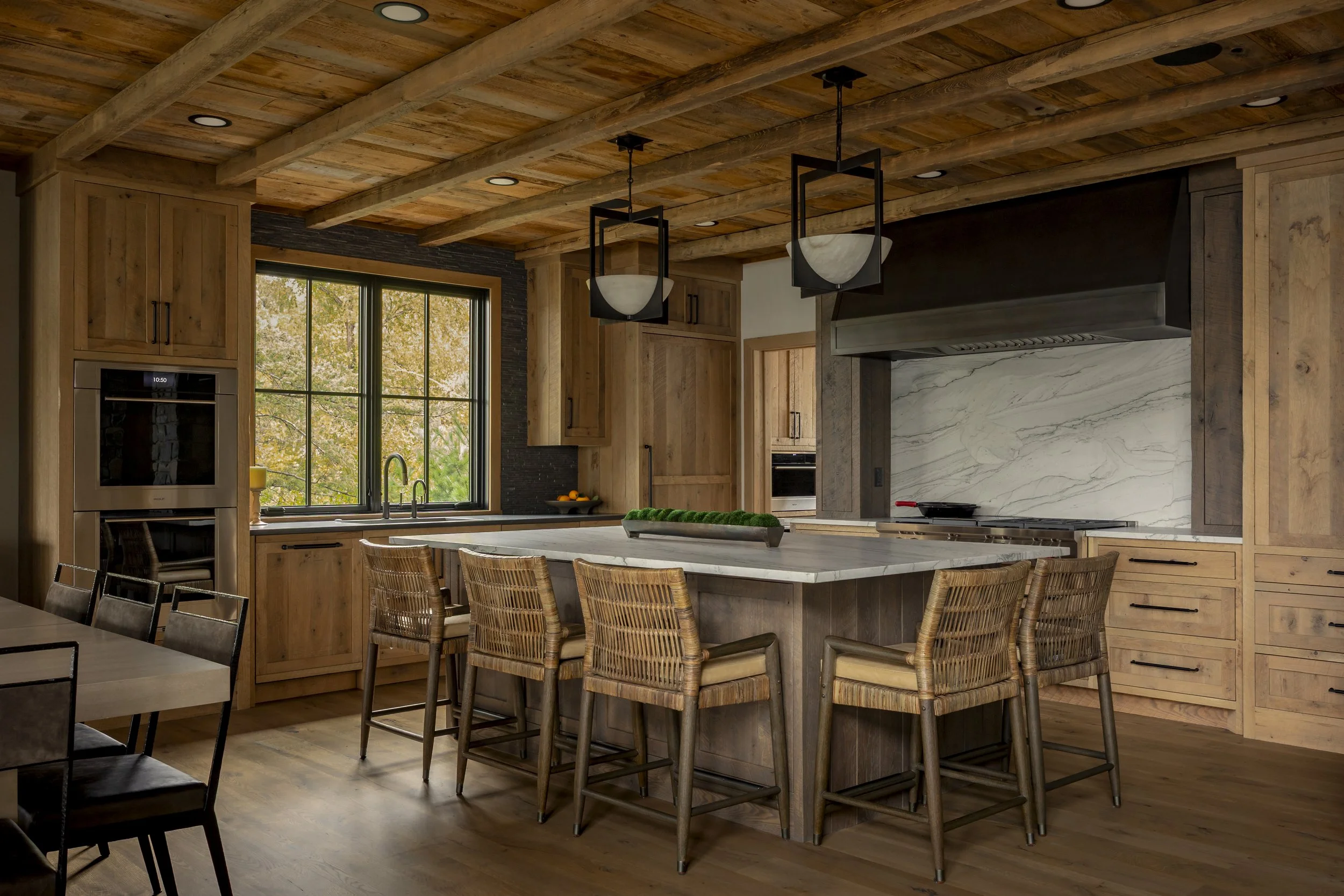Modern kitchen with wooden cabinets, a large island with seating, black pendant lights, a marble backsplash, and a window with a view of trees.