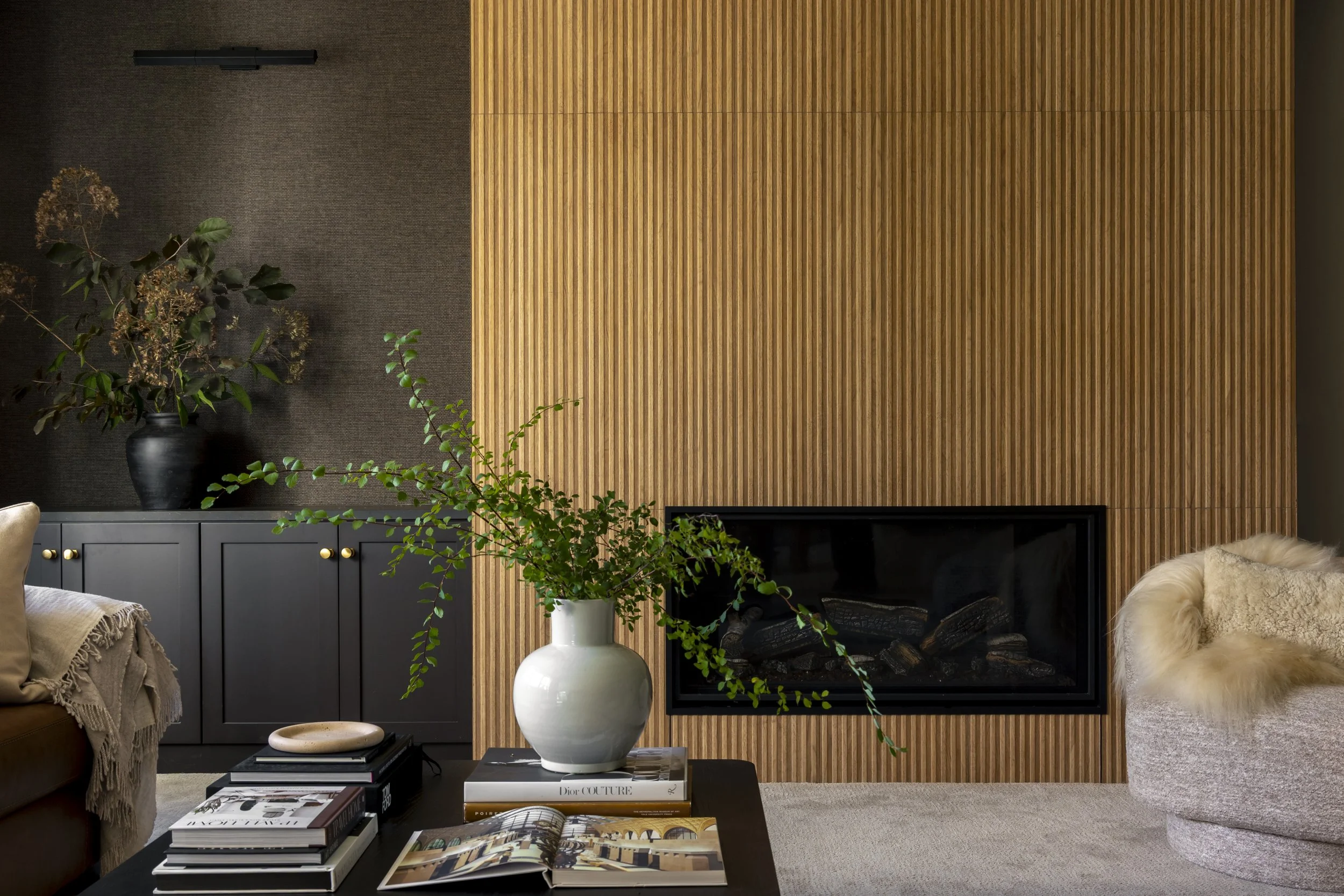 Modern living room with a fireplace, wooden panel wall, gray cabinet, plants, stacks of books, and a plush armchair.