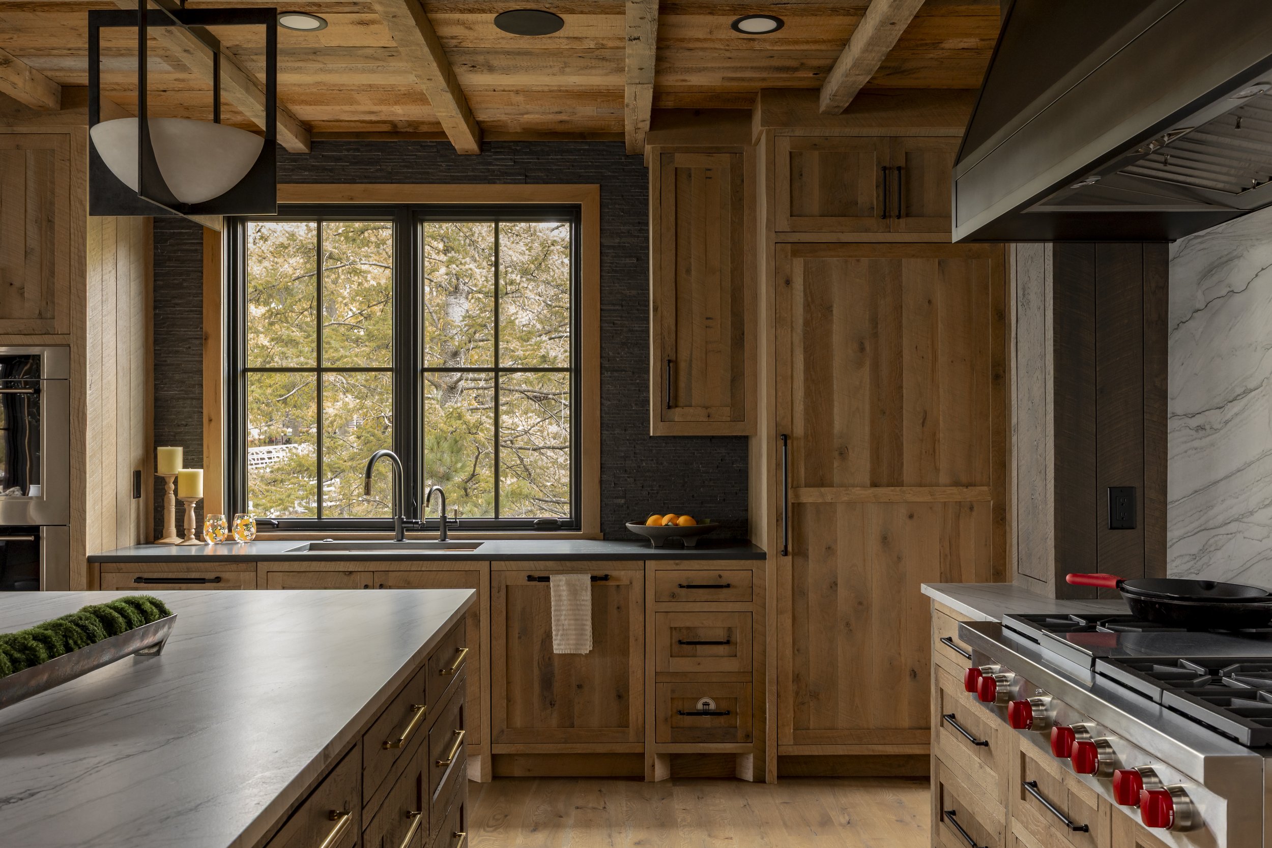 Rustic kitchen with wooden cabinets, marble countertops, a large window with a view of trees, and stainless steel appliances.