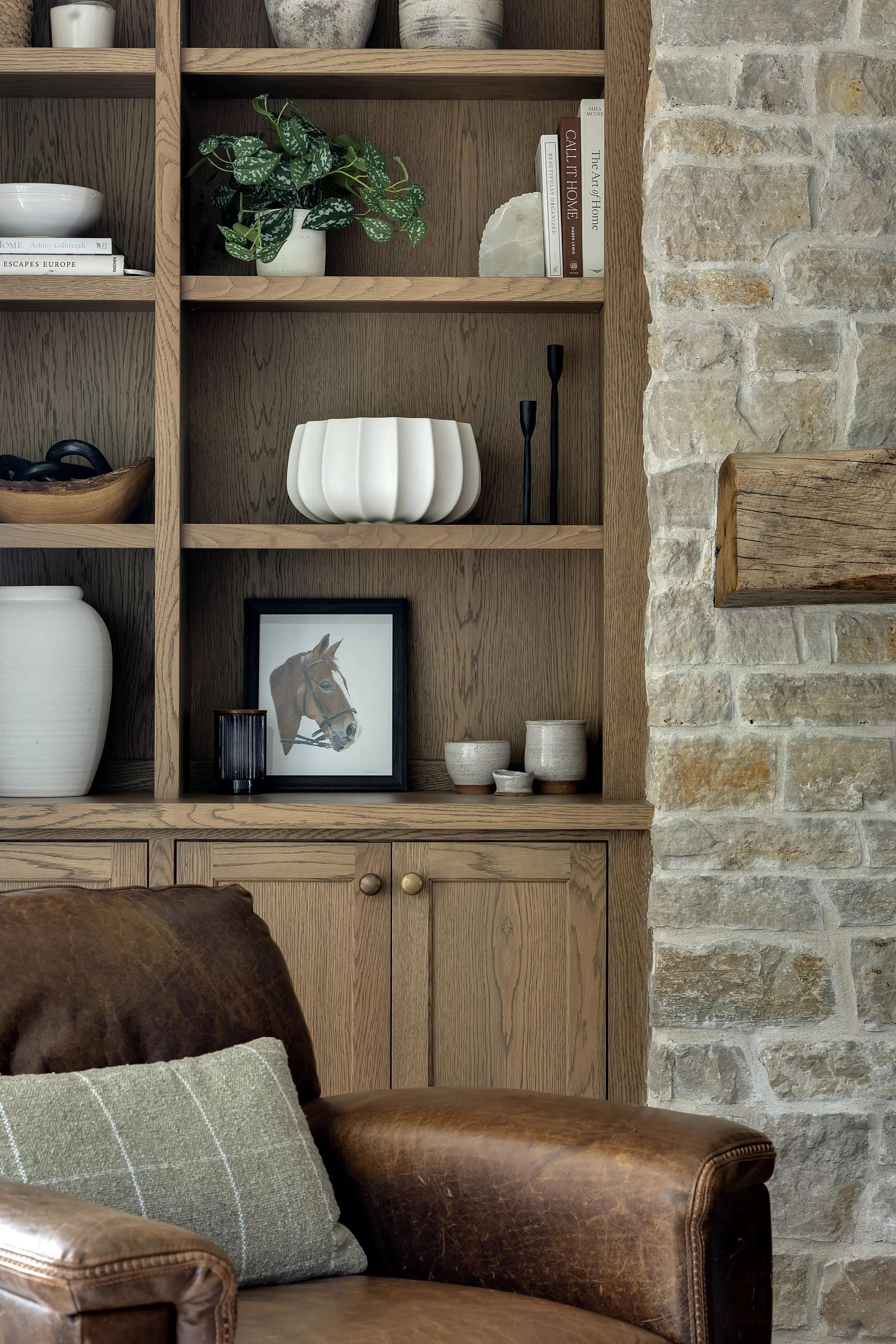 A wooden bookshelf with various decor items, a framed horse portrait, some books, and a small plant, set against an exposed brick wall in a cozy living room.