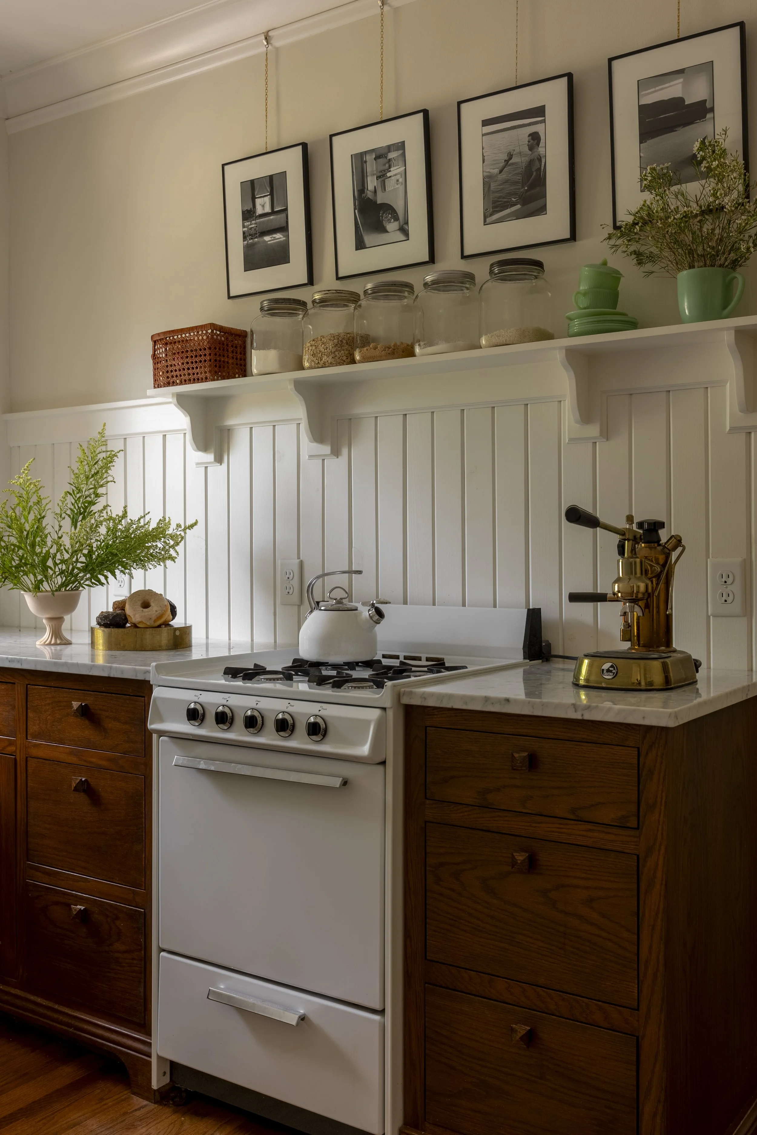 A cozy kitchen corner featuring a white stove with a white kettle on top, wooden cabinets, a marble countertop, and a white backsplash. There are green plants, vintage jars, framed black-and-white photos, and kitchen accessories on a shelf above.