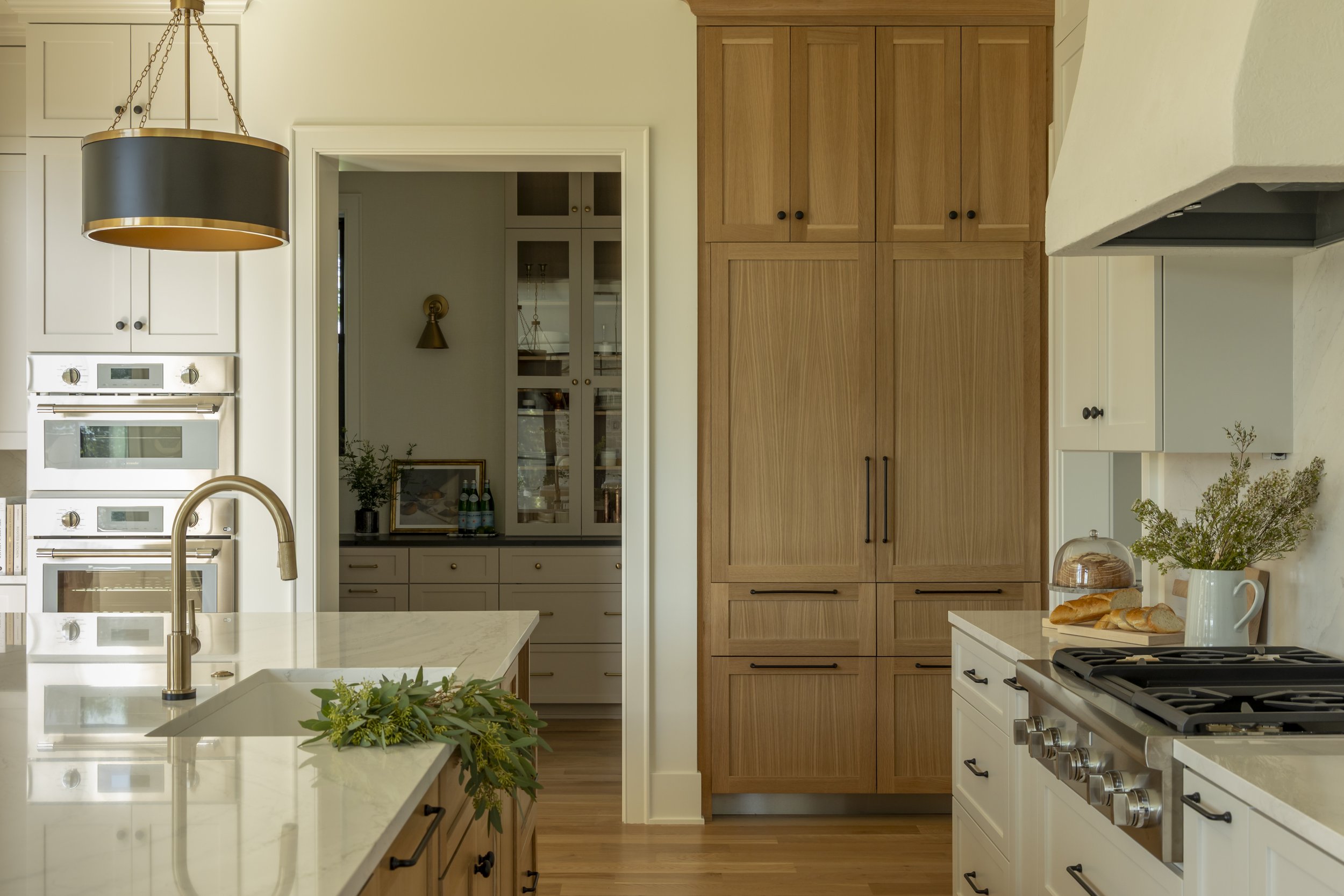 Modern kitchen with white cabinets, wooden pantry, marble countertops, stainless steel oven, and a center island with greenery. There is a black and gold hanging light fixture above the island.