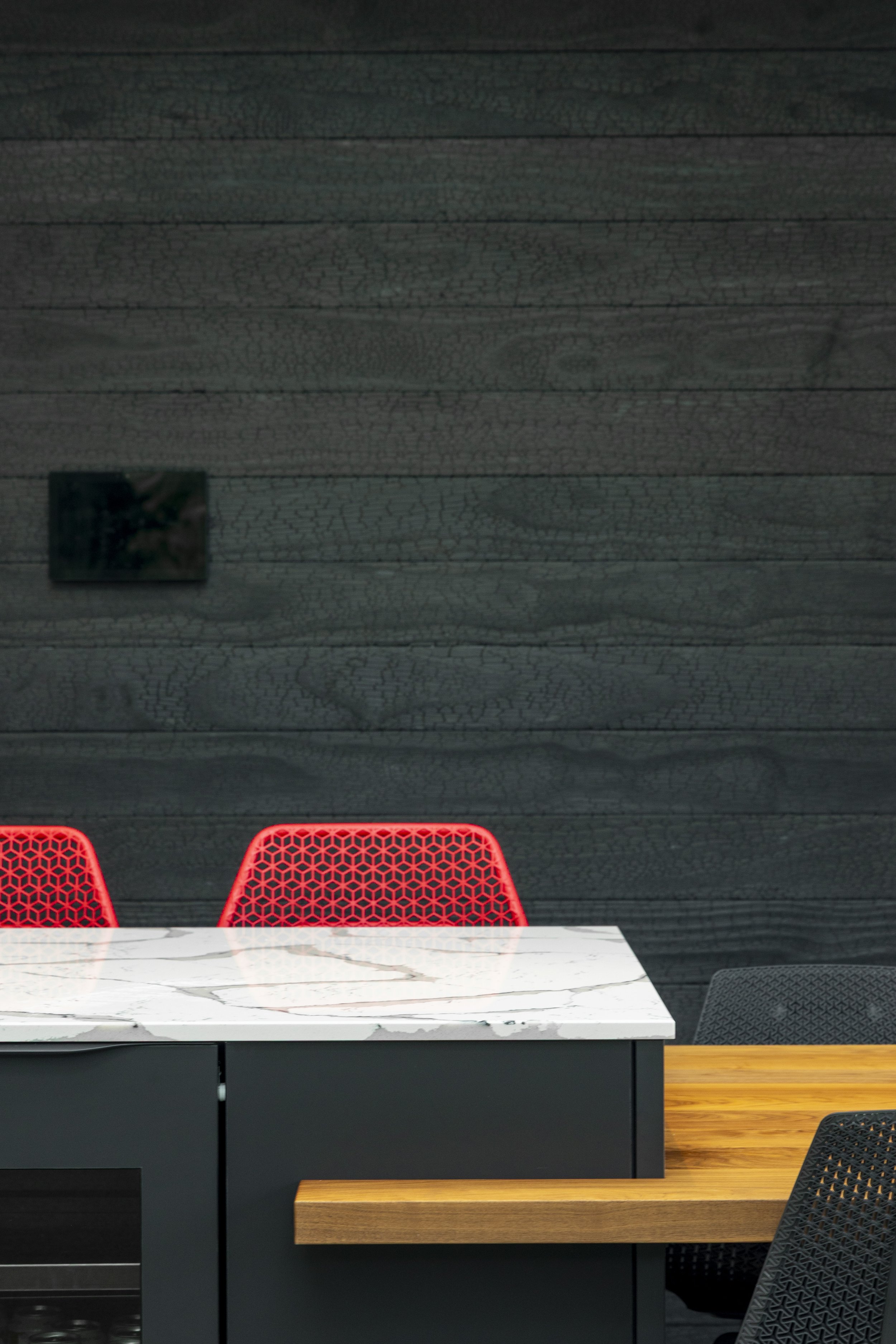 Part of a modern dining or meeting area with a black wall featuring a textured, cracked paint finish, a white marble-topped table, and red chairs with geometric cutouts.