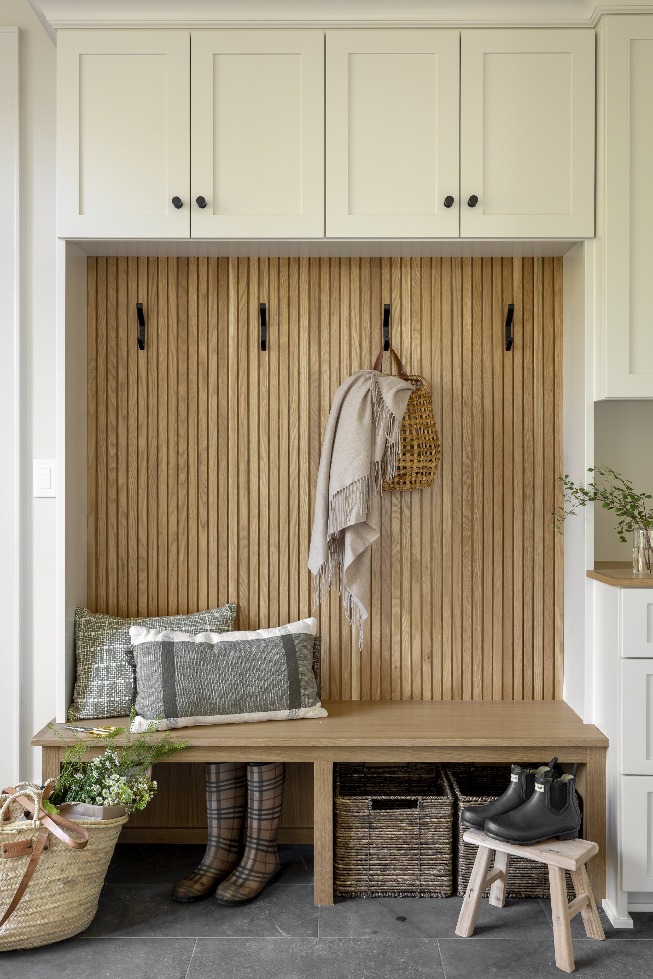 Entryway with wooden bench, pillows, rain boots, woven baskets, and coat hooks on a wood-paneled wall.