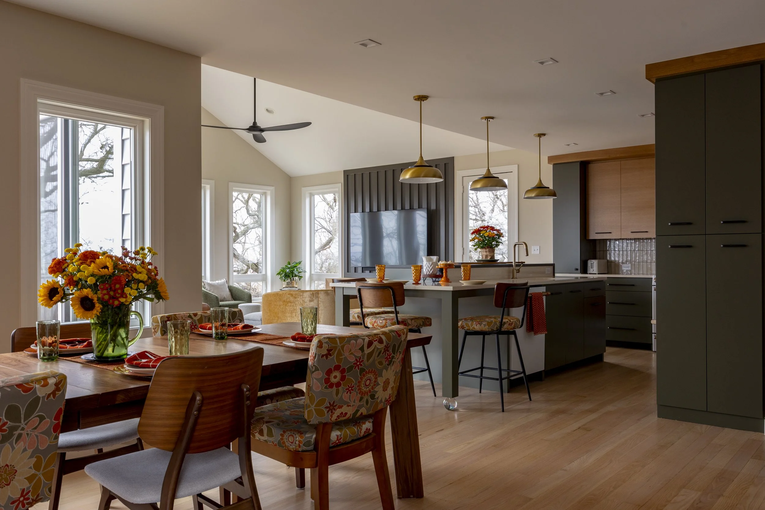 Open-concept living space with a dining area, kitchen, and sitting nook with large windows showing leafless trees outside, decorated with flowers and modern furniture.