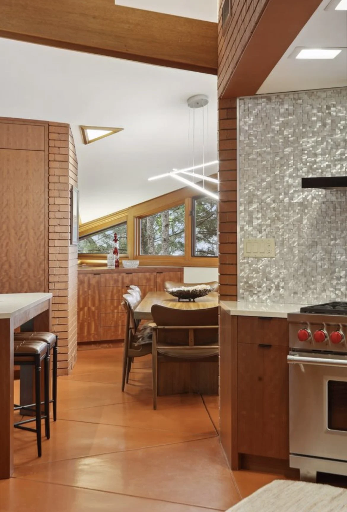 Interior view of a kitchen and dining area with wood and brick accents, a sloped window with a wooded outdoor scene, modern lighting fixtures, and a stove.