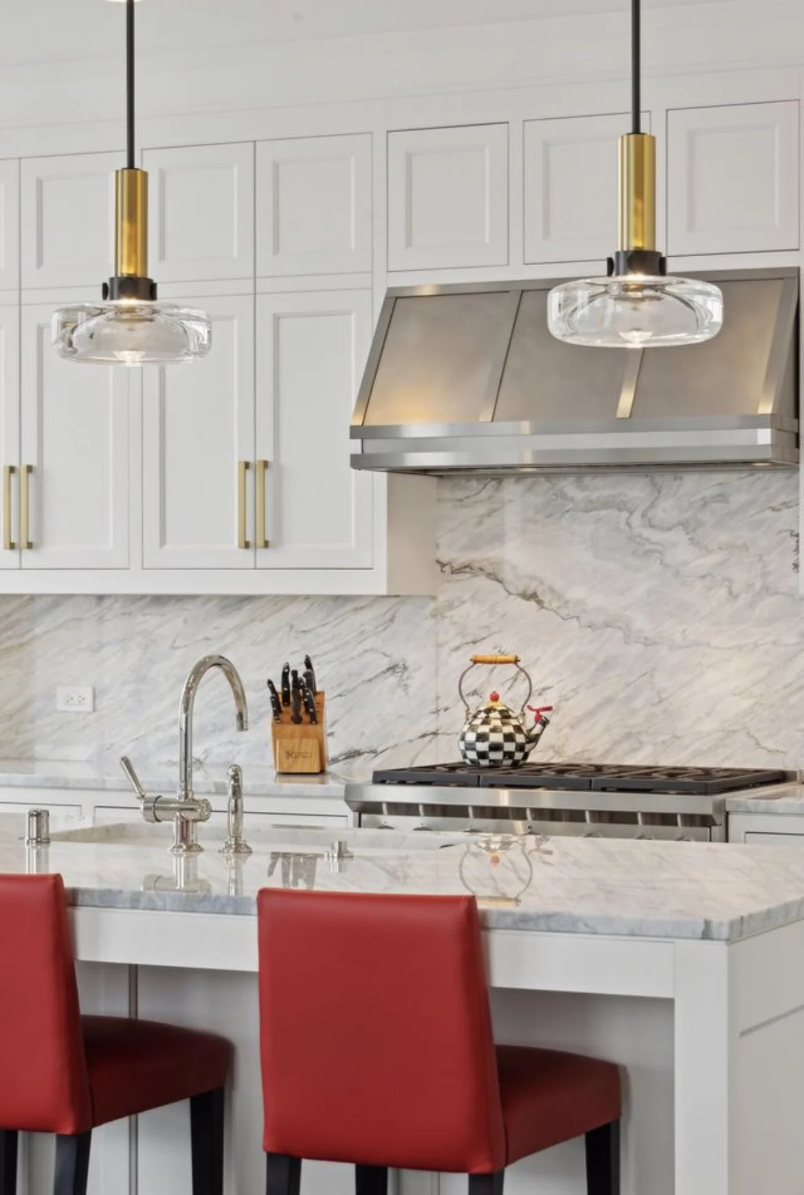 Modern kitchen with white cabinets, white marble countertops, and backsplash. Two pendant lights hanging above the counter, a stainless steel range hood over a stove, a teapot on the stove, a knife block on the counter, and three red chairs at the ki