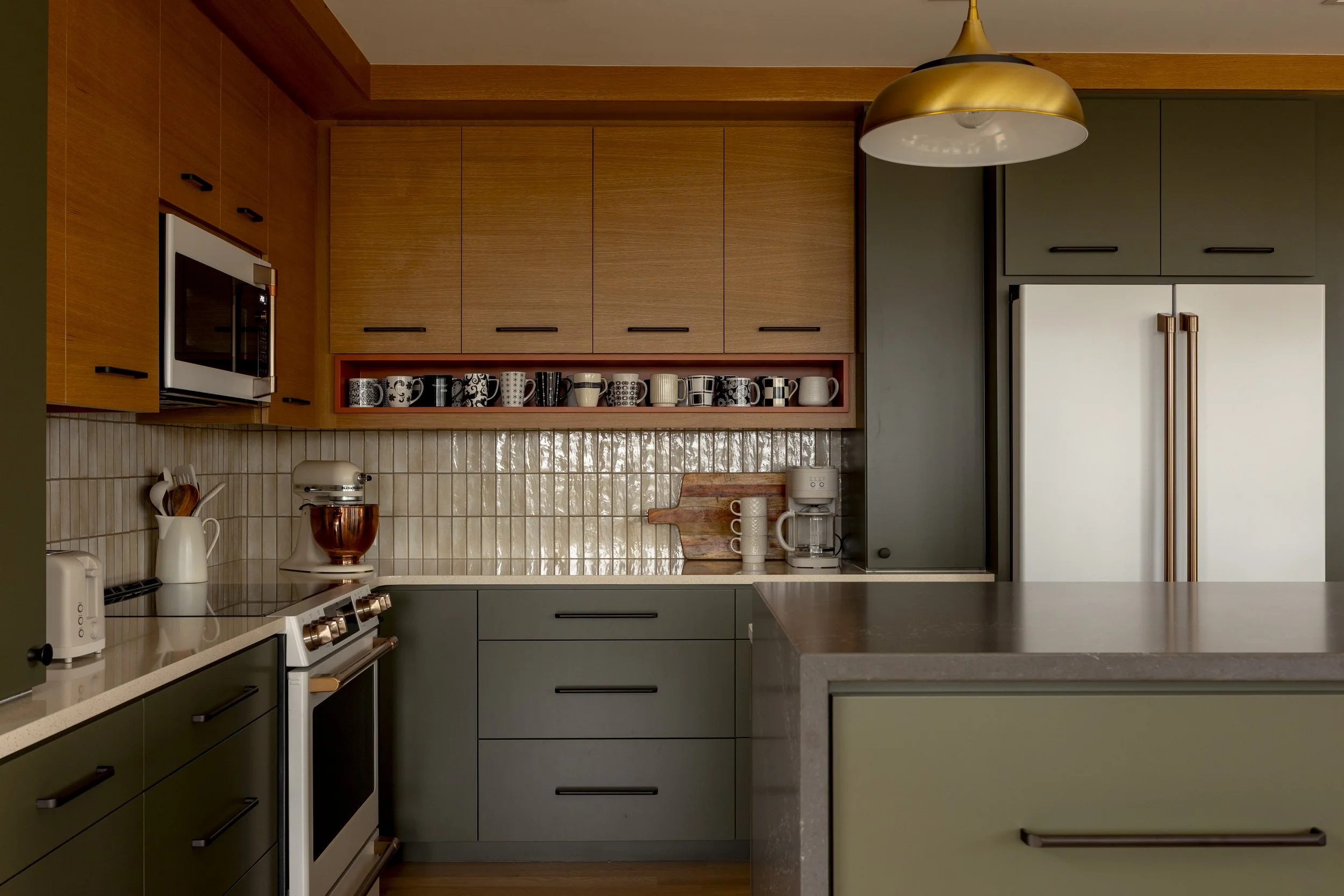 Modern kitchen with green lower cabinets, wood upper cabinets, a gray refrigerator, a cream countertop, and a gold pendant light, with various kitchen appliances and decorative mugs.