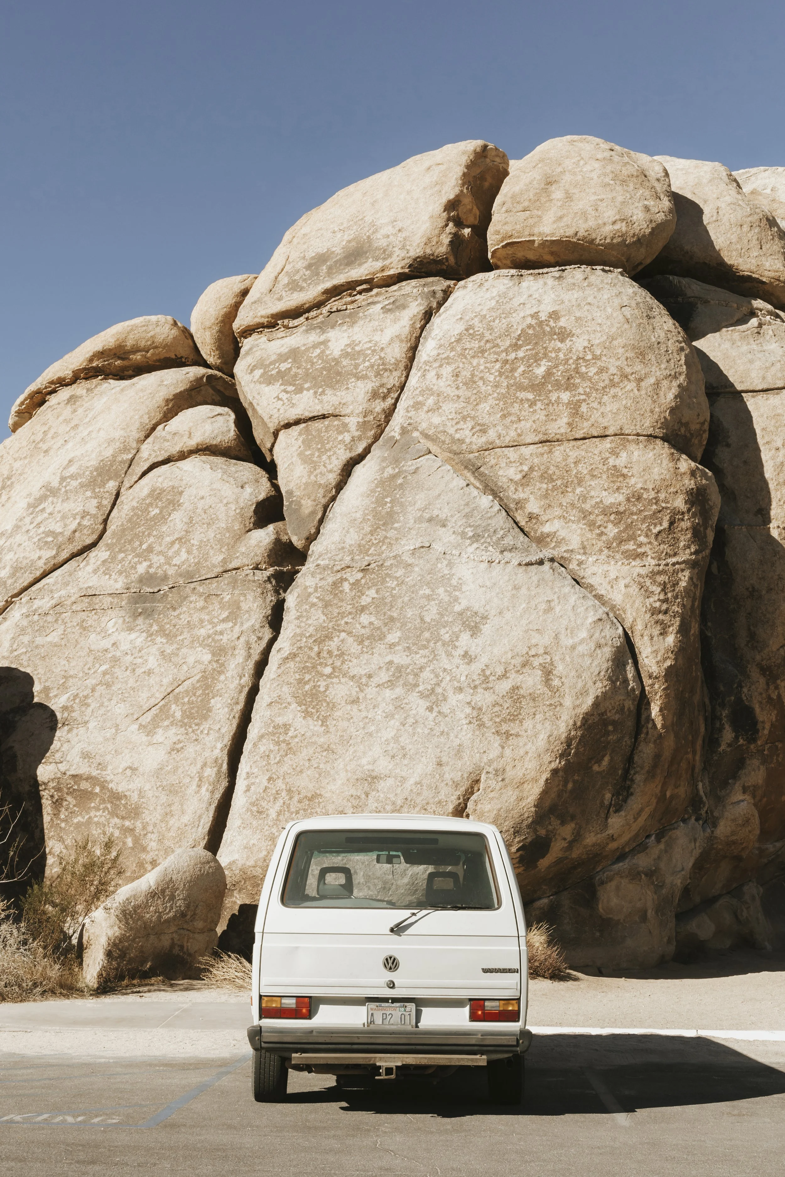 A white Volkswagen Vanagon parked in front of large desert rocks under a clear blue sky.