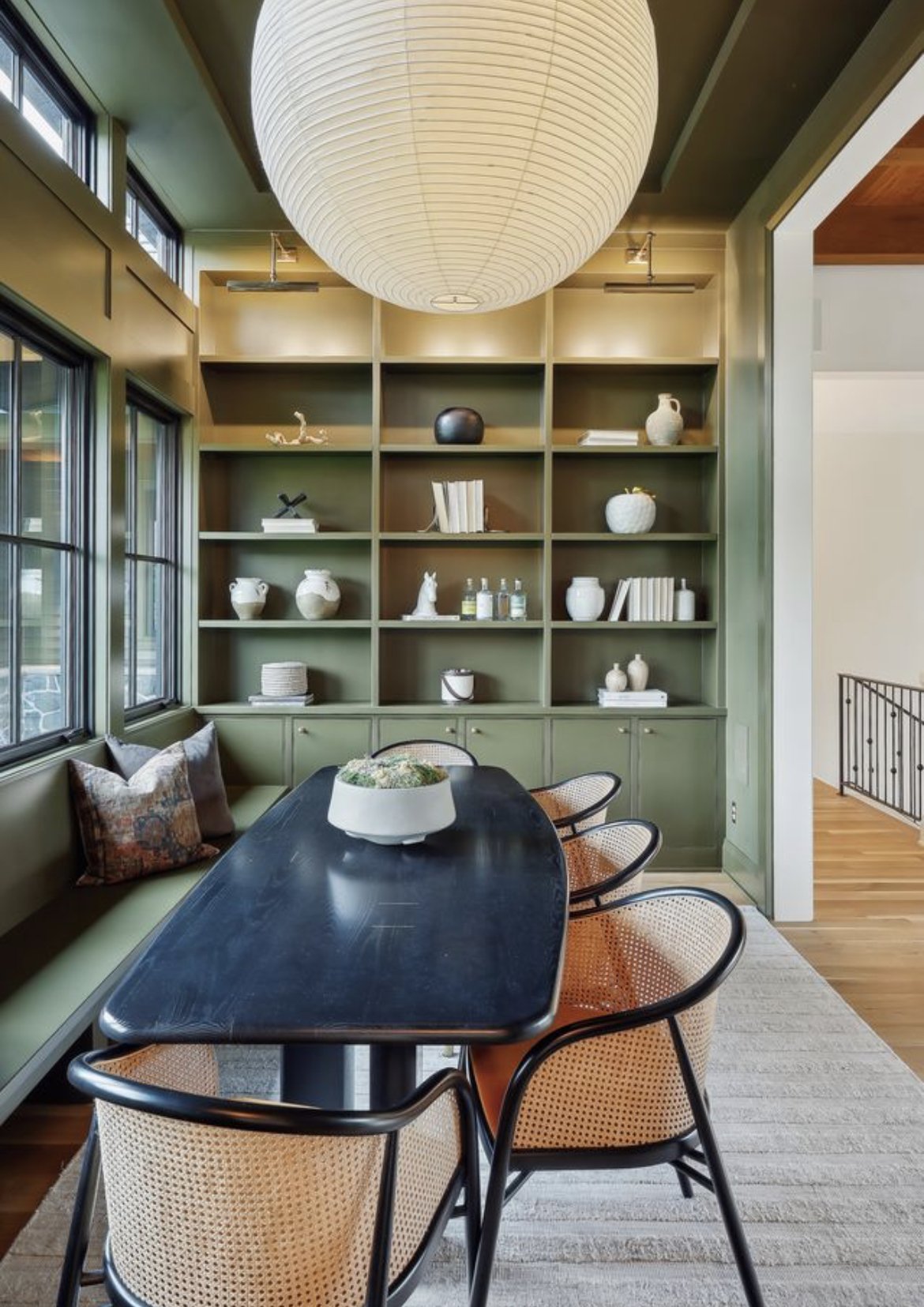 A dining room with a black oval table, four rattan chairs, a built-in green bench with pillows, large windows, a green bookshelf with decorative objects, and a large round paper lantern ceiling light.