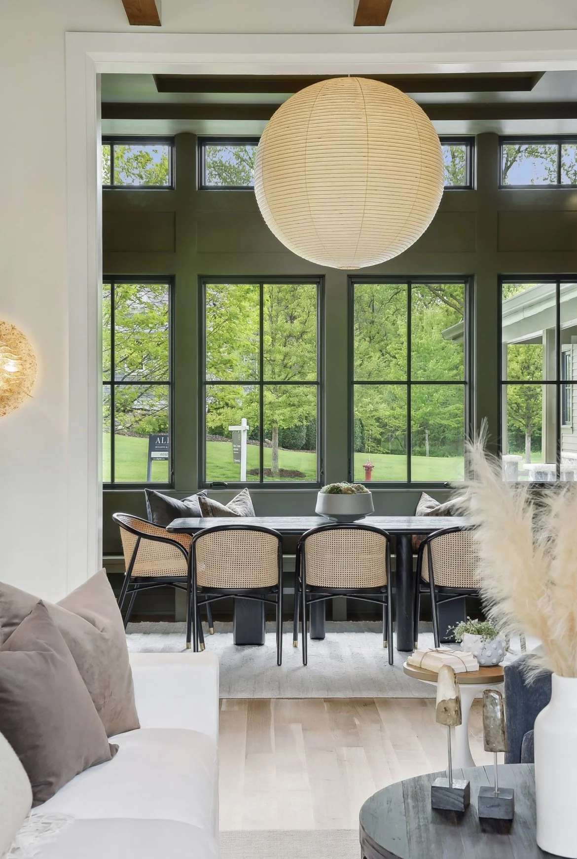 A cozy dining room with a black rectangular table, six beige and black chairs, large windows with a view of greenery, a large round paper lantern hanging from the ceiling, and stylish decorative items on a side table.