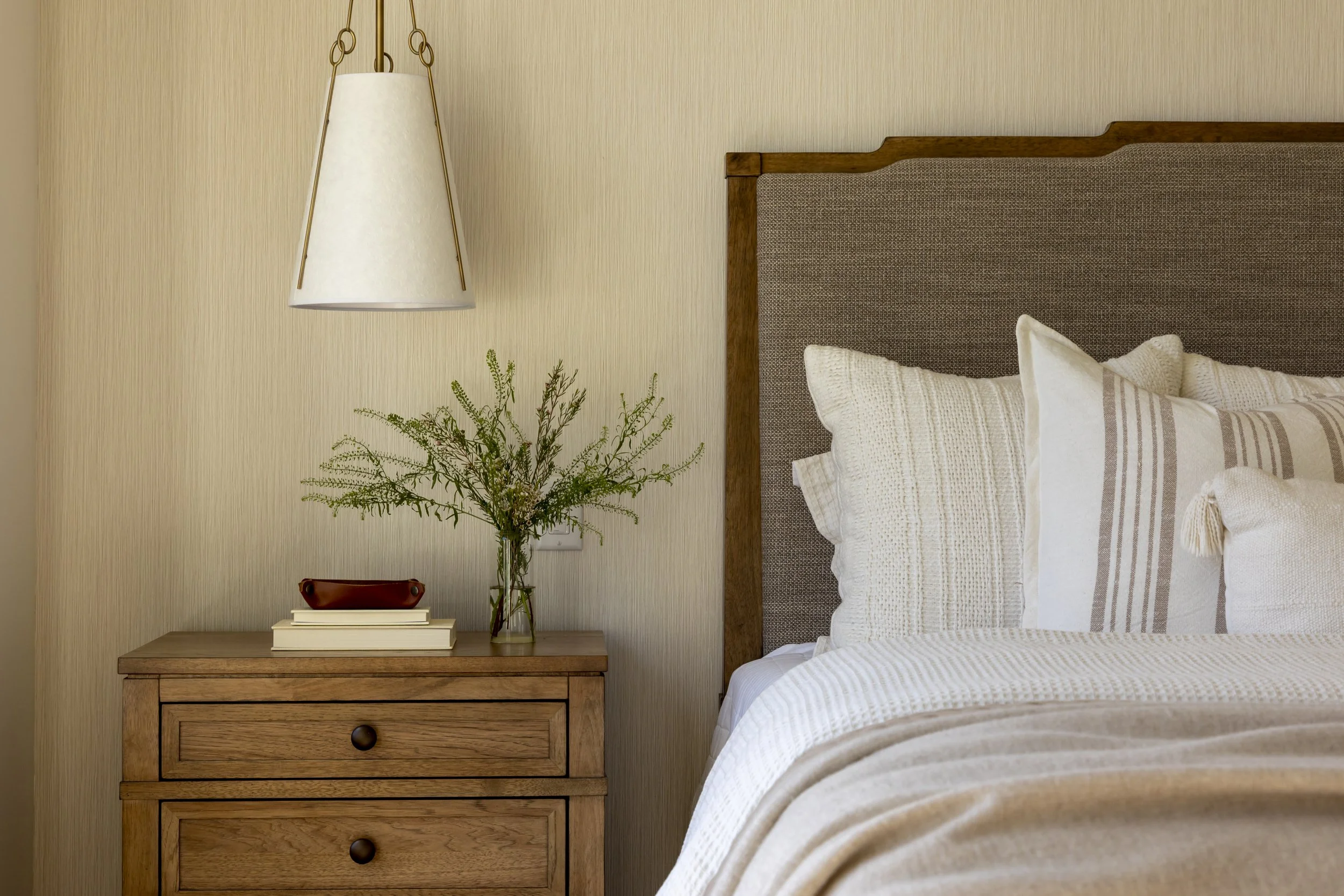 A cozy bedroom corner featuring a wooden bedside table with a glass vase of greenery, a stack of books, and a ceramic dish. Above, a hanging white lampshade with gold accents. Part of a bed with white and beige linens and a padded fabric headboard.