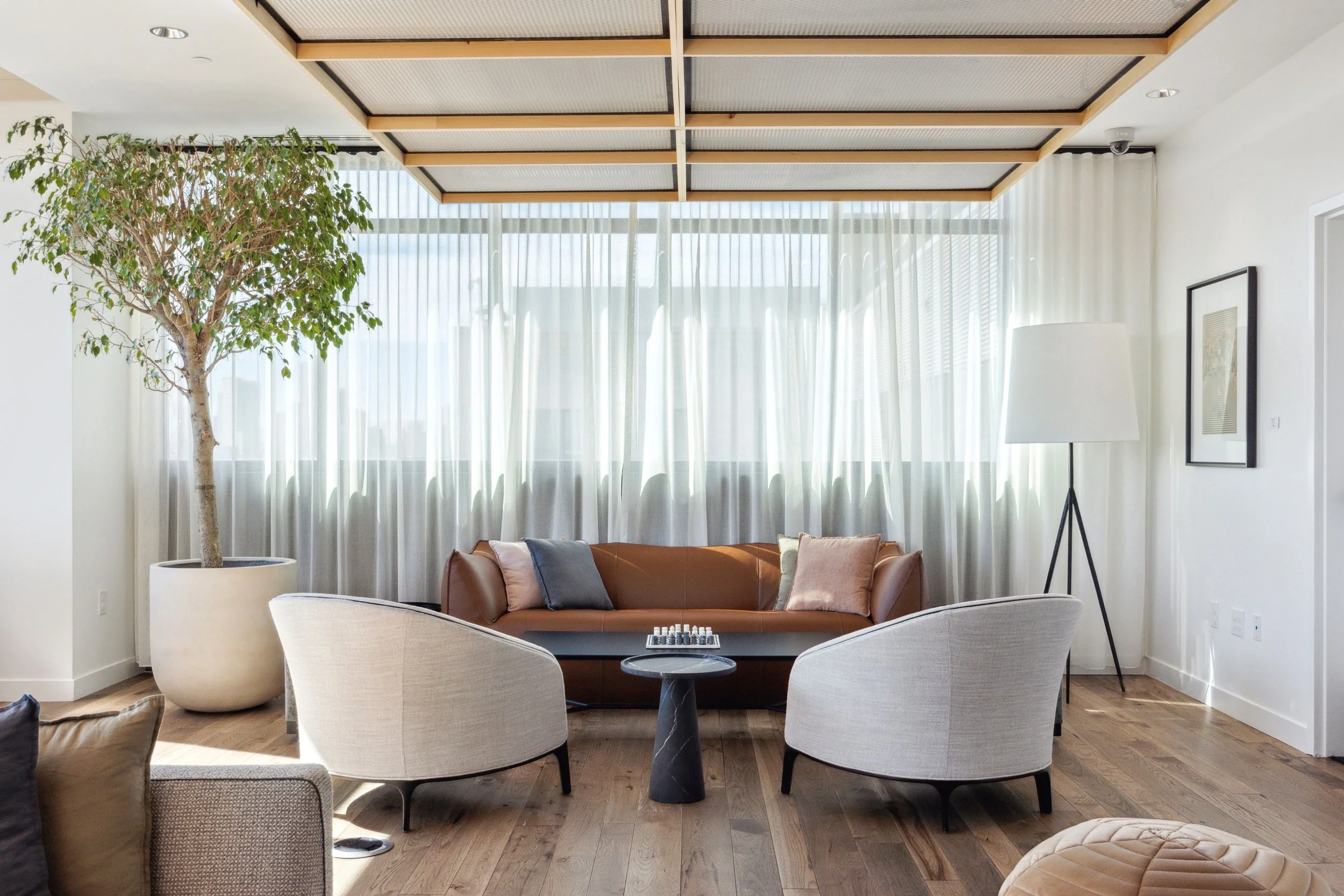 Living room with large window, sheer curtains, a potted tree, a brown leather sofa with pillows, two white armchairs, a black round side table with chess, and a white floor lamp, hardwood floor, framed artwork on wall.