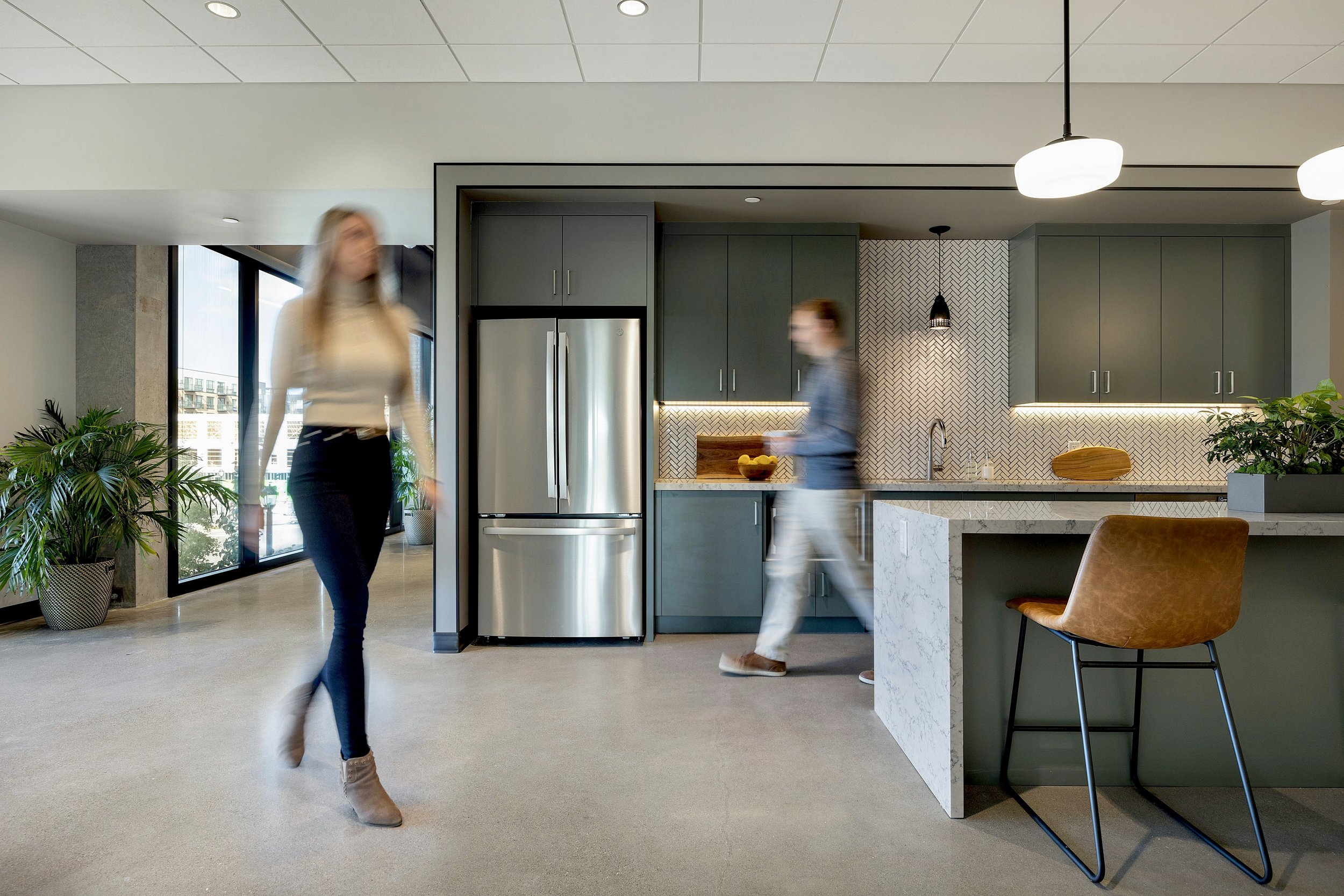 Modern kitchen with two women walking through, blurred from movement. Bright space with large windows, green cabinets, stainless steel refrigerator, marble countertops, and decorative backsplash. Potted plants and a brown stool near the counter.