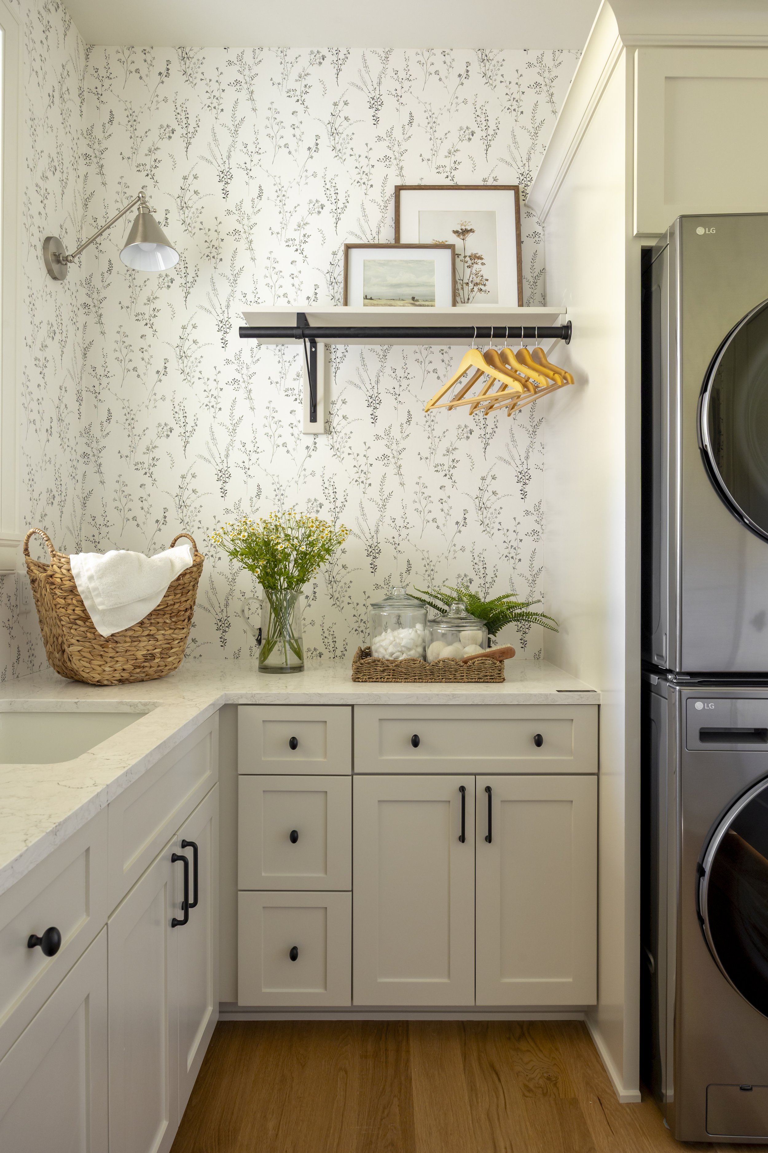 Laundry room with white cabinetry, a marble countertop, a woven basket with a towel, glasses with flowers, jars with cotton balls, framed artwork on a shelf, and a stainless steel washer and dryer.